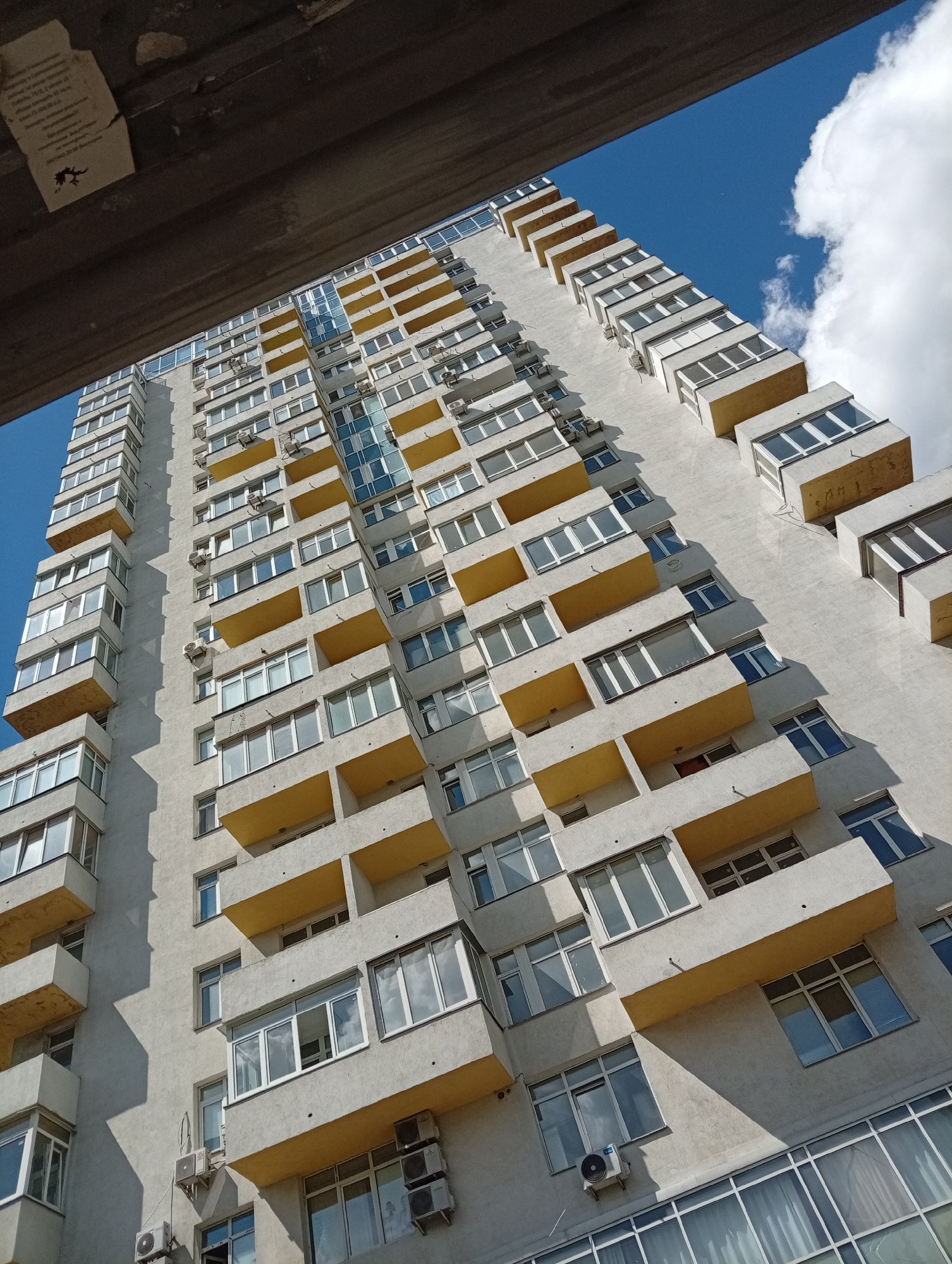 This media file shows a close-up, low-angle shot of a tall, modern apartment building under a bright blue sky with white clouds. The building is primarily light gray with yellow balconies. Numerous windows, some with white trim and others with blue reflections, punctuate the facade. Several air conditioning units are visible mounted on the exterior walls. A dark, textured overhang is present in the upper portion of the frame, casting a shadow.
The perspective is from below, looking upwards at the building, which dominates the frame. The sun appears to be shining brightly, creating distinct shadows that highlight the architectural details of the balconies and windows. The time of day is likely daytime, given the bright sunlight and clear sky. The weather appears to be fair and sunny. There is no specific location information discernible from the image itself. No people are visible in the media file.
On the overhang in the upper left corner, there is a piece of paper with some text written on it. The text is in a dark color and appears to be in a non-Latin script, possibly Cyrillic. Below the text, there is a small black graphic of a creature that resembles a lizard or dragon.