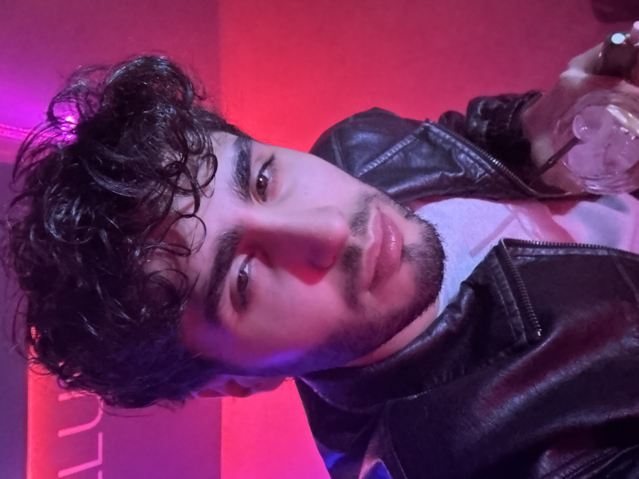 A male individual, appearing to be in his late 20s to early 30s, is depicted in a close-up, waist-up shot, looking directly at the camera. He has dark, curly hair, pronounced eyebrows, light brown eyes, and a trimmed beard with a mustache. He is wearing a black leather jacket over a light-colored t-shirt. His right hand is visible, holding a clear glass containing ice cubes and a transparent liquid, with a dark-colored straw. A ring is present on his right pinky finger. The background is dimly lit and features strong pink and purple neon lighting, casting a glow on the subject. A partial neon sign, displaying the letters "LU," is visible in the upper left background. The environment suggests an indoor nightlife venue, such as a club or lounge, located in the city of Yerevan, Armenia.