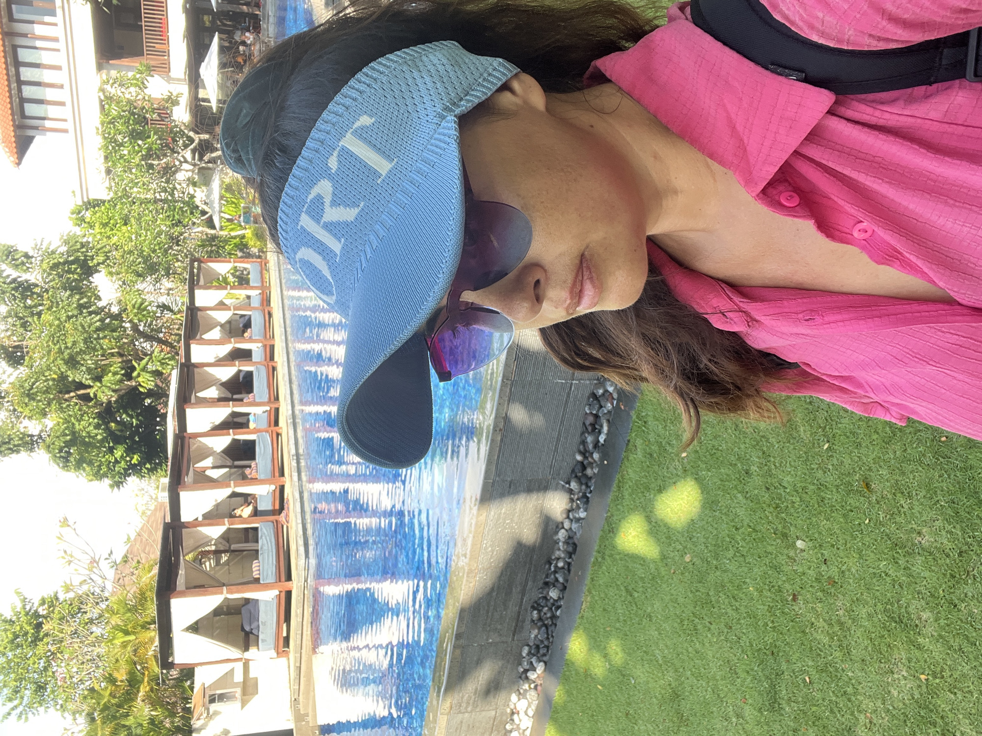 The image depicts a woman wearing a pink collared shirt and a light blue visor with the word "SPORT" on it, standing in front of a swimming pool. She is also wearing sunglasses. Behind her are several cabanas with white curtains, a swimming pool with blue water, and trees. The setting appears to be a resort or hotel in Kuta Selatan, Indonesia, characterized by its tropical vegetation and resort-style architecture. The sun is shining, casting shadows on the grass. The woman is looking to her right, away from the camera. The overall scene suggests a vacation or leisure setting.
