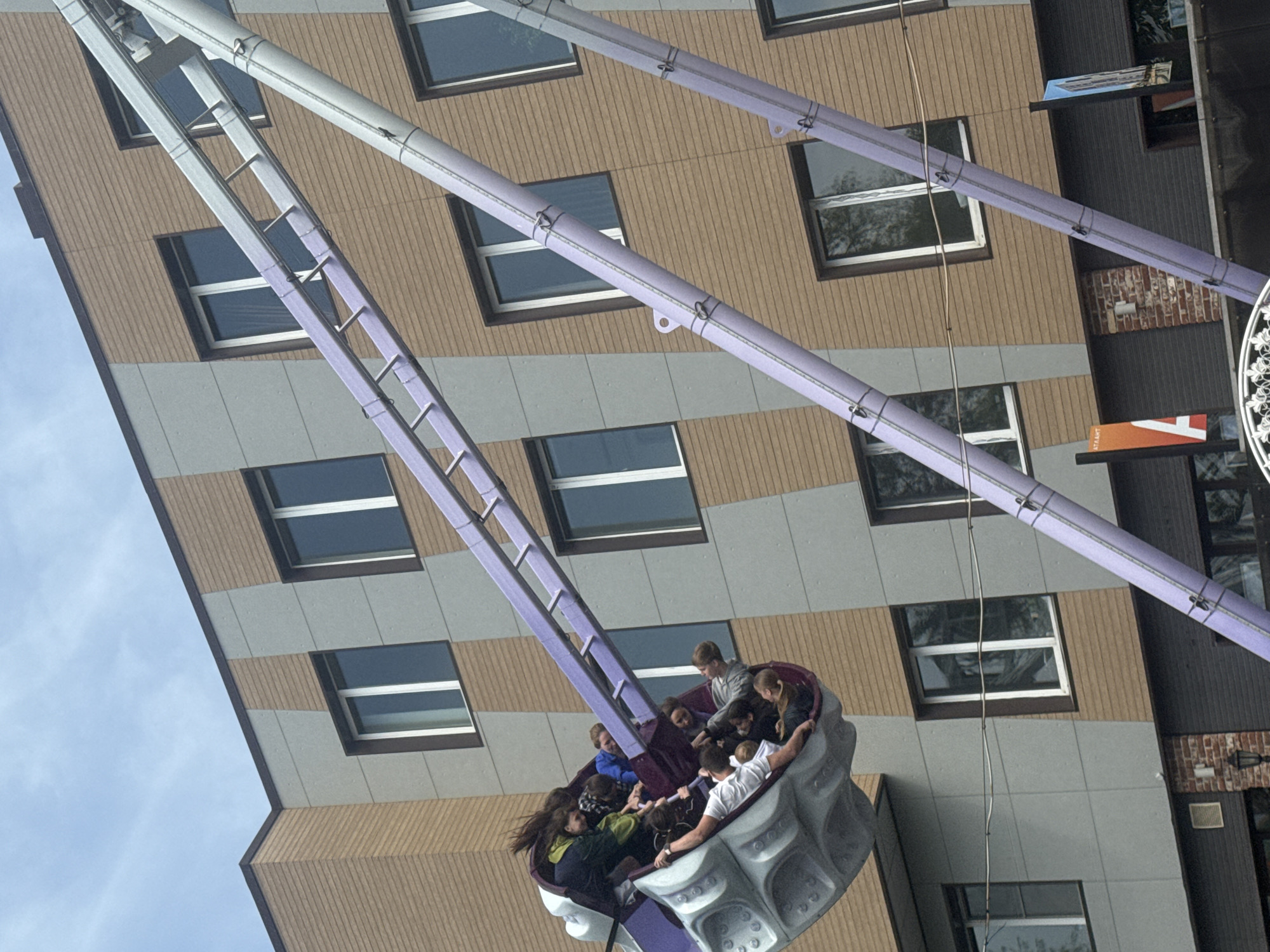 The image depicts a fairground ride, likely a type of pendulum or rotating arm ride, in front of a multi-story building. A group of people, mostly appearing to be teenagers or young adults, are seated in the gondola of the ride. The building has a façade of alternating gray and brown horizontal panels, with windows visible on each story. The ride structure itself is predominantly light purple or lavender. The sky is visible in the background, suggesting an outdoor location. The location is identified as Vladivostok, Russia.