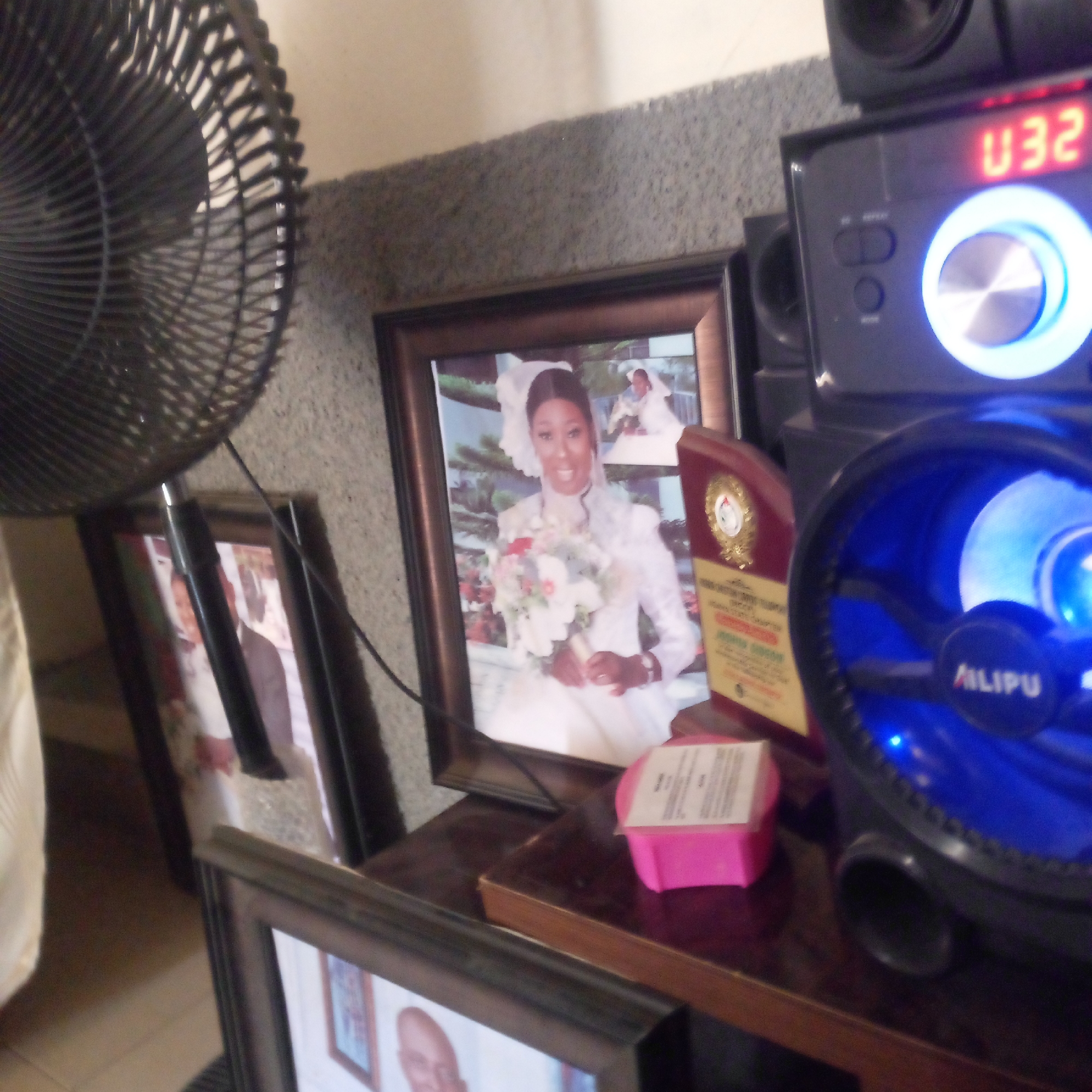 The image shows a close-up of a collection of framed photographs and electronic devices resting on a dark wooden surface. A black electric fan with a protective grille is positioned on the left, partially obscuring the view. Behind the fan, several framed photographs are arranged, the most prominent featuring a woman in a white wedding dress holding a bouquet. Another smaller framed photo and a wooden plaque are also visible. To the right of the photos is a black electronic sound system with speakers, illuminated by blue and red lights, and the word "AILIPU" is visible on the device. The background is a grey textured wall.