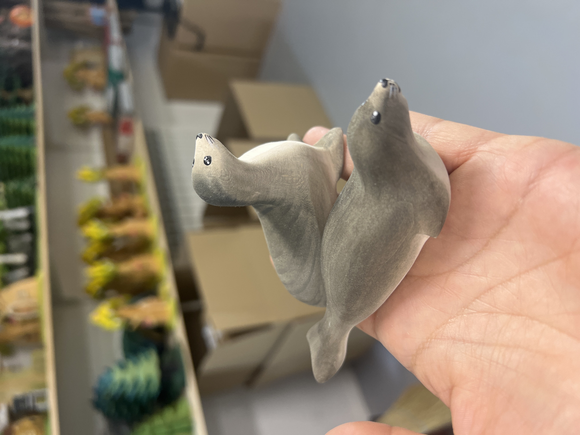 Two grey, wooden figurines of seals are held in a person's hand. The seals are smooth and have black, circular eyes and small black noses. The upper seal is perched on top of the lower seal, and both are positioned with their heads looking upwards and to the right. The background of the image is out of focus but appears to be an indoor setting with shelves holding various colorful objects, possibly figurines or crafts, and several cardboard boxes. The scene is brightly lit.