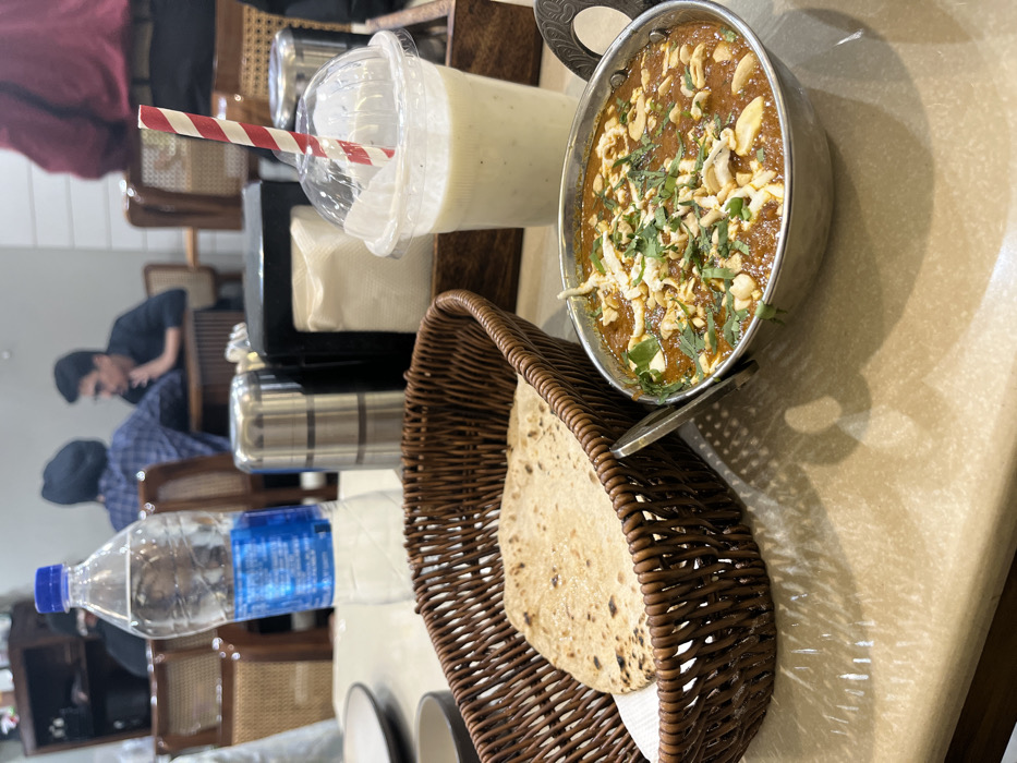 The image shows a table setting in a restaurant. The table is covered with a beige tablecloth and has a brown wooden surface. There is a wicker basket on the table containing a round flatbread. A bowl of curry, garnished with parsley, is to the right of the basket. A glass of milk with a red and white striped straw sits to the right of the bowl. A bottle of water sits at the left of the basket. The background shows a partial view of other tables and chairs, as well as a person in red seated at a table and two people sitting at a table in the distance. The restaurant appears to be in Sahibzada Ajit Singh Nagar, Punjab, India.