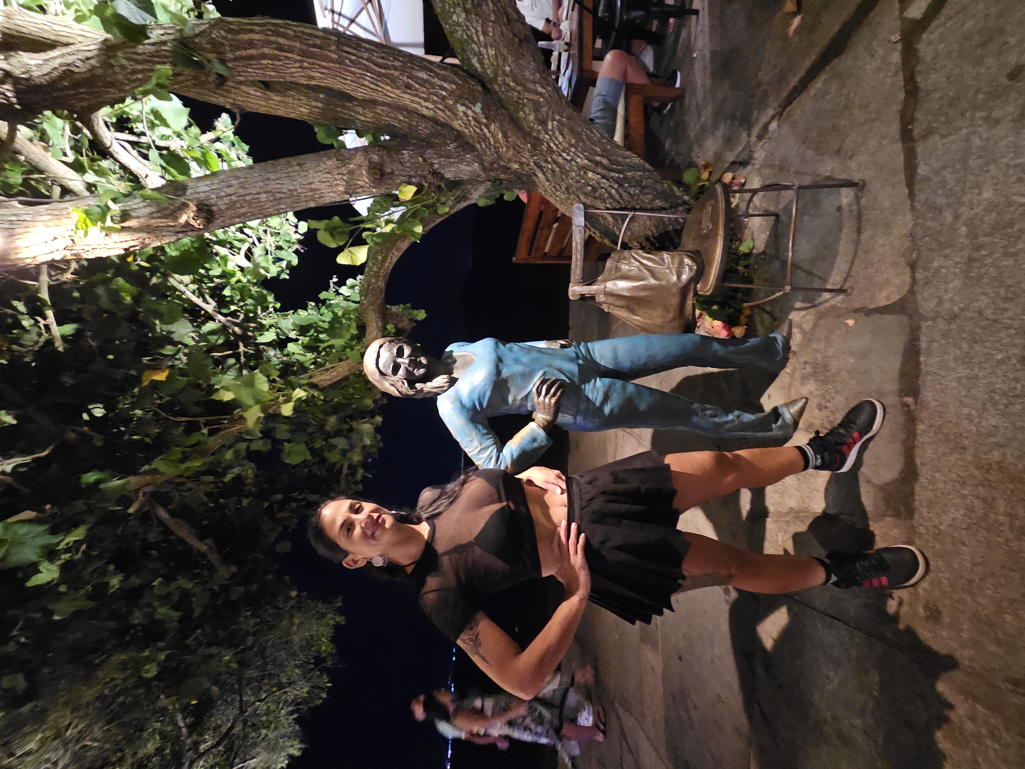 A woman in a black outfit poses for a photo with a blue, skeletal statue under a tree. The scene is outside at night. The location is likely Armação dos Búzios, Brazil. The woman is fair-skinned and has dark hair pulled back from her face. She is wearing a black top with a mesh overlay, a short black skirt, and black sneakers with red accents. The statue is lying on its back, and the woman is positioned next to it. A tree is behind the statue. There's a metal chair and a gold-colored bag behind the statue. In the background, there are hints of other people and lights. The ground is concrete, and there's some form of lighting.