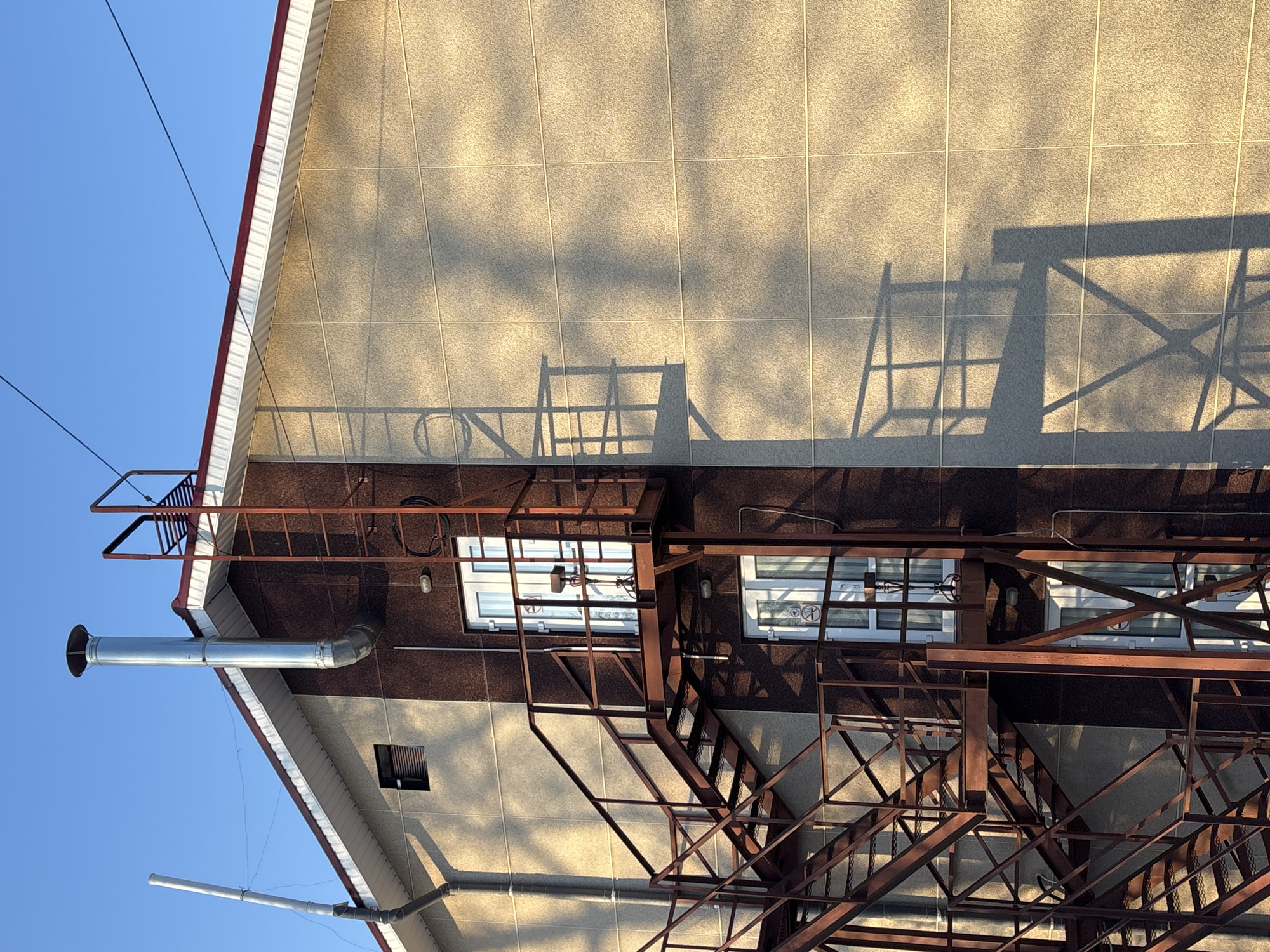 The image shows a building in Chaikovsky, Russia. The facade is divided into a light tan upper section with a tiled appearance and a dark brown lower section. A metal fire escape structure is attached to the building, casting shadows on the upper facade. Two windows with white frames are visible, along with a small ventilation window. A metal chimney extends from the side of the building. The sky is clear blue.