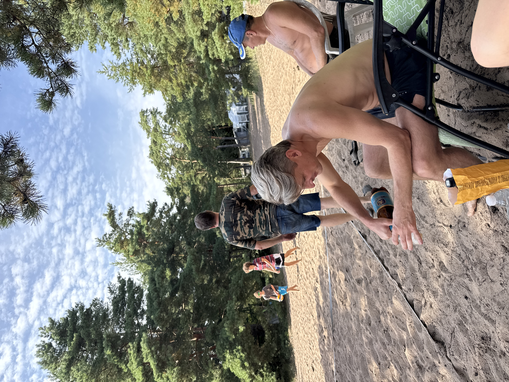 The image depicts an outdoor scene on a sandy beach, likely in Zelenaya Roshcha, Russia, with a background of pine trees and a blue sky with scattered clouds. In the foreground, a shirtless man with blondish-grey hair is seated in a black folding chair, pouring from a brown bottle into a clear cup. He is wearing dark shorts. To his right, another shirtless man with a blue baseball cap is seated in a white folding chair. Further in the background, two young boys are playing on the sand. One boy, wearing blue shorts and sunglasses, holds a yellow ball. Another boy, wearing a striped shirt and dark shorts, is running. A man in a camouflage shirt and denim shorts stands watching them. Behind the people, there are some tents and structures, suggesting a campground or a beach area with amenities. The sand is a light brown color and has tire tracks visible. A thin black line is laid across the sand in the foreground. A yellow bag and a white water bottle are on the sand near the seated man.