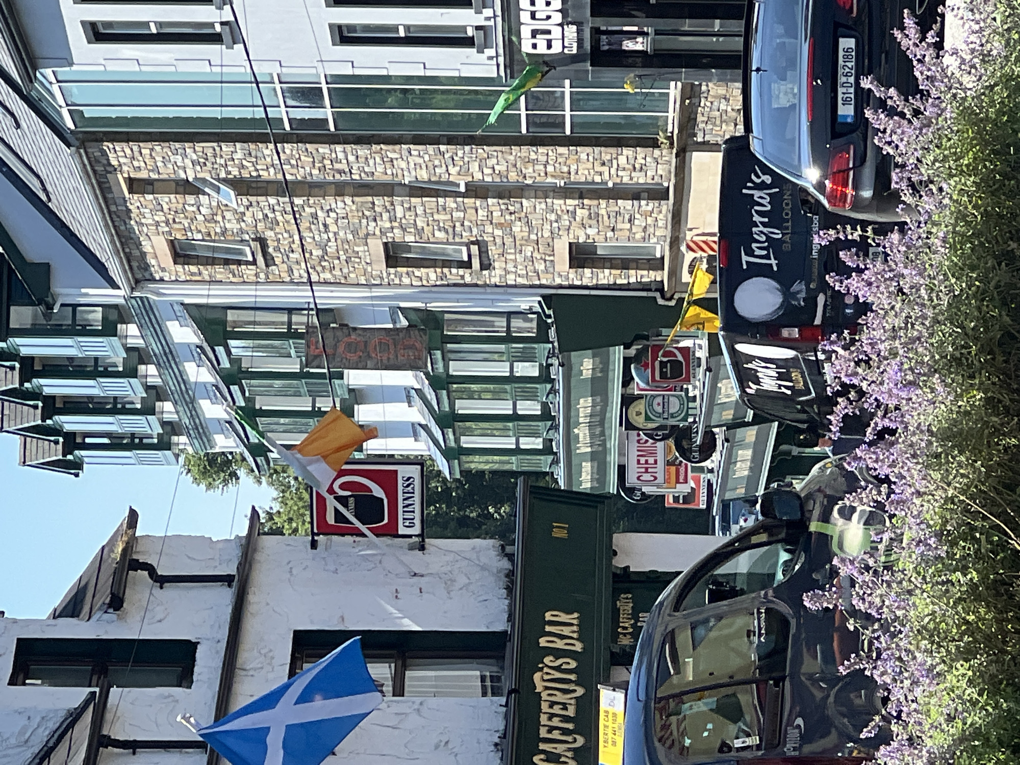 The image shows a street scene in Donegal, Ireland, on a sunny day. In the foreground, there are flowering purple plants. Behind the plants, several vehicles are visible, including a dark-colored car with a license plate that reads "161-D-62186" and a black van with "Ingrid's BALLOONS" written on its side. Further back, a green awning with "CAFFERTY'S BAR" in gold lettering spans across the front of a building. Above the awning, a sign for Guinness is displayed. Two flags are visible: a Scottish flag (blue with a white saltire) and an Irish flag (green, white, and orange tricolor). The buildings in the background appear to be a mix of stone and white-plastered facades, with a classic European architectural style featuring bay windows and tiled roofs. The overall impression is of a bustling town center.