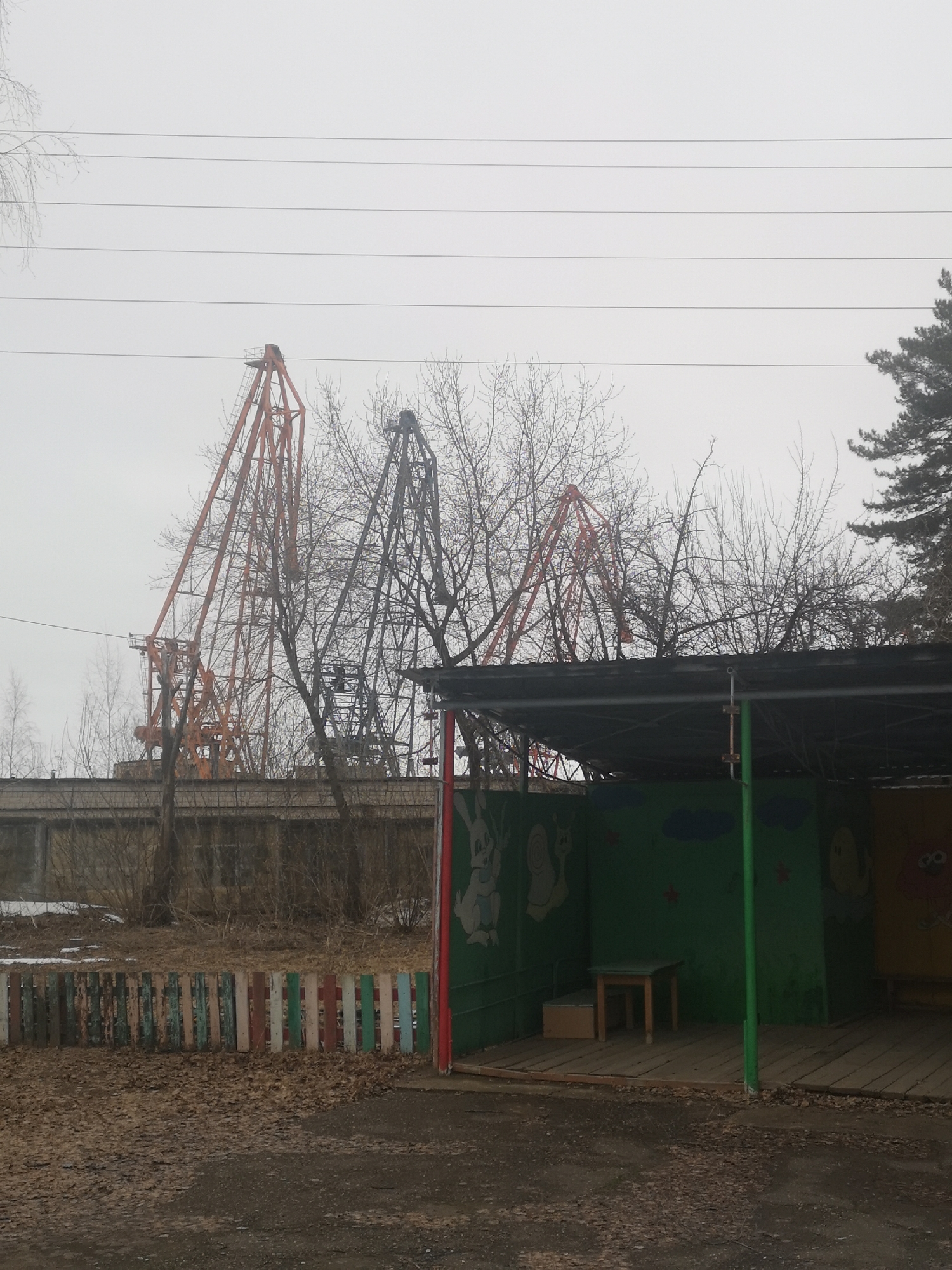 The image depicts a playground in what appears to be an industrial area in Chaikovsky, Russia. In the foreground, there's a dilapidated playground structure with painted cartoon figures on the walls. A small wooden table and bench are inside the structure. Outside, a colorful, damaged fence borders the playground. The ground is covered in leaves and debris. In the background, several industrial cranes dominate the skyline, partially obscured by bare trees and a gray, overcast sky. Power lines run horizontally across the upper part of the image. The overall impression is one of neglect and urban decay in an industrial setting. No people are visible in the image.