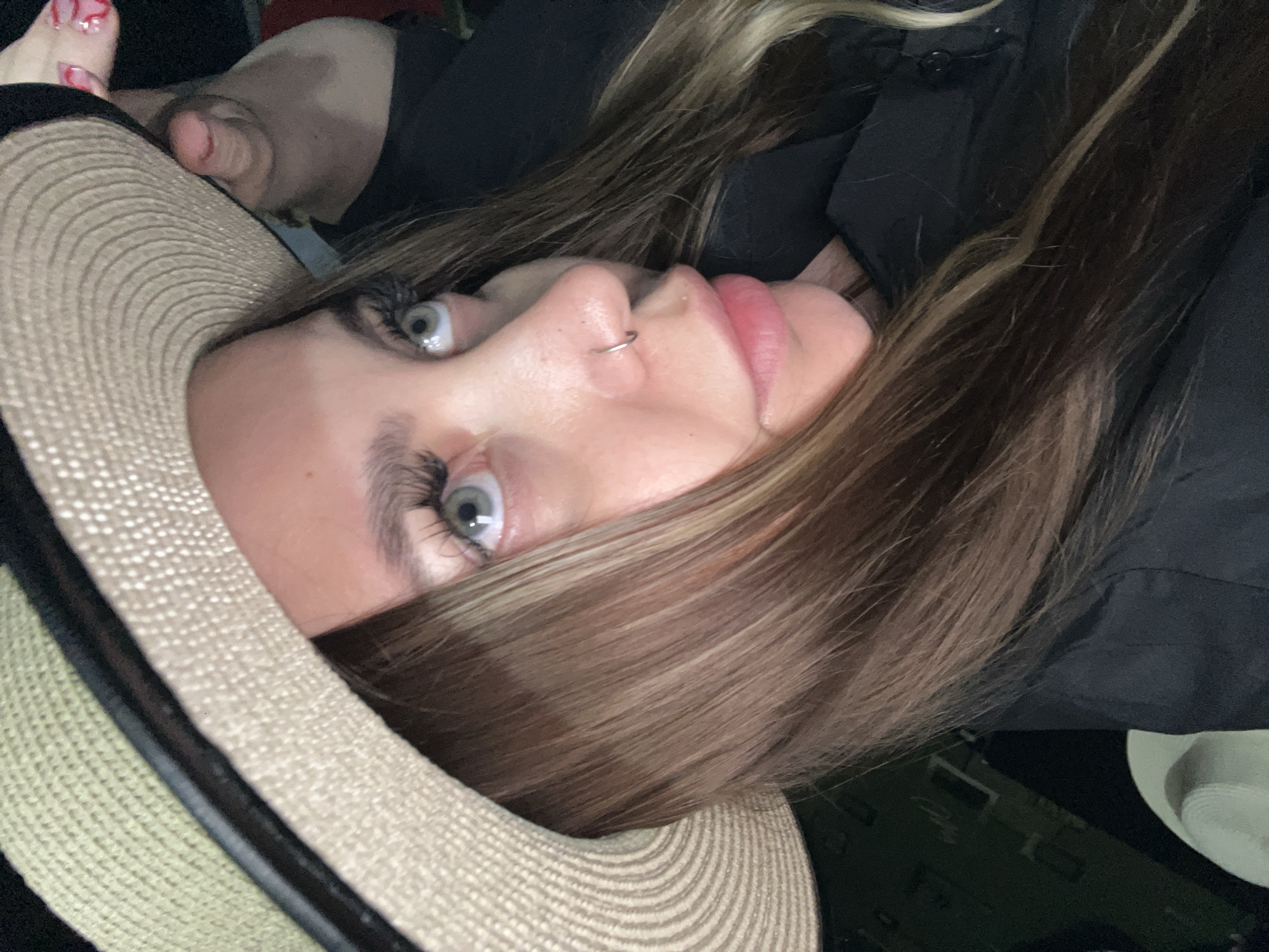 A close-up, tilted portrait shows a young woman wearing a sun hat. Her blonde-highlighted hair is partially visible. She has light skin, a nose ring, and long eyelashes. Her eyes are focused off-camera. The hat is woven with a dark band. Toes with painted nails are visible at the top left. The woman is wearing a black shirt or jacket. The background is dark. There's no evidence to suggest this image was taken in Novopokrovskaya, Russia.