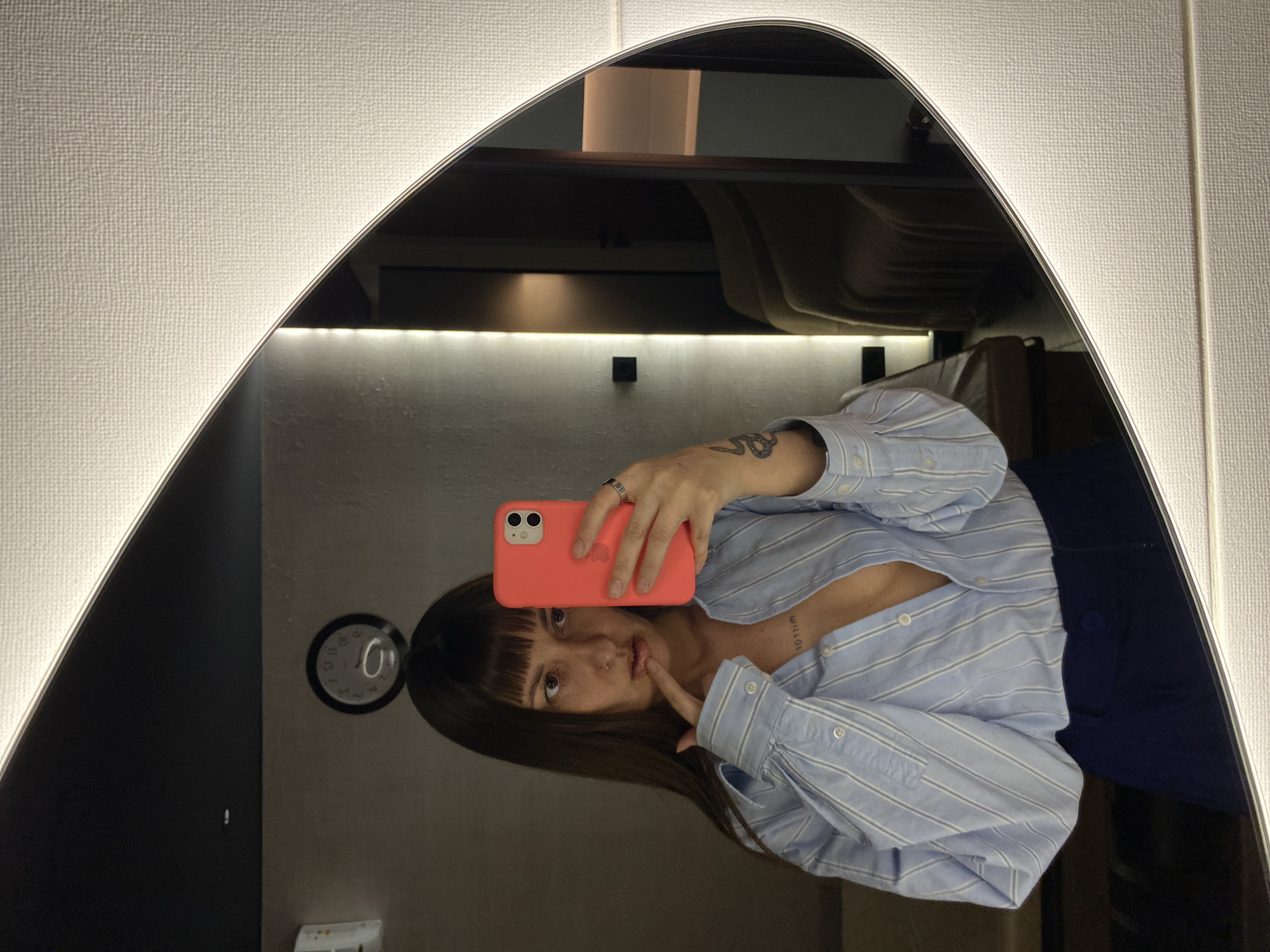 A mirror selfie shows a woman holding a coral-colored smartphone in front of her face. She has brown hair with bangs, wears a light blue striped shirt, and a dark blue garment. Her left index finger is touching her lower lip. Visible in the background of the mirror's reflection is a clock on the wall and elements of a room with white textured walls. The location of the image is not discernible as being in Votkinsk, Russia.