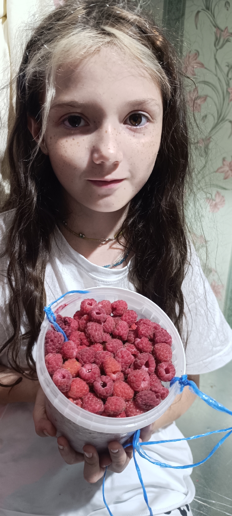 In this close-up, vertical medium shot, a young girl with long, dark hair and noticeable freckles on her face holds a bucket overflowing with ripe raspberries. Her hair has light streaks, and she wears a white t-shirt and a beaded necklace. The background features a floral-patterned wallpaper. The girl is positioned in the foreground, looking directly at the camera, with the bucket of raspberries held in front of her chest. Blue strings are tied to the sides of the bucket. The lighting appears to be natural and diffused. The image does not provide any visual information that allows for the identification of a specific location within the city of Karsha, Russia.