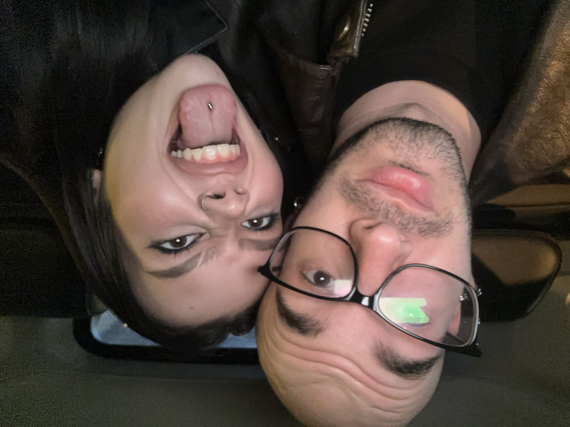 Two people are in the image, which has been rotated 180 degrees. On the left, a woman with dark hair is sticking her tongue out, revealing a tongue piercing. She also has a nose ring and visible eye makeup. On the right, a man with a beard and glasses looks into the camera. The location is unclear, and there is no evidence to suggest it is Moscow, Russia. They are both wearing dark clothing and appear to be posing for a selfie.