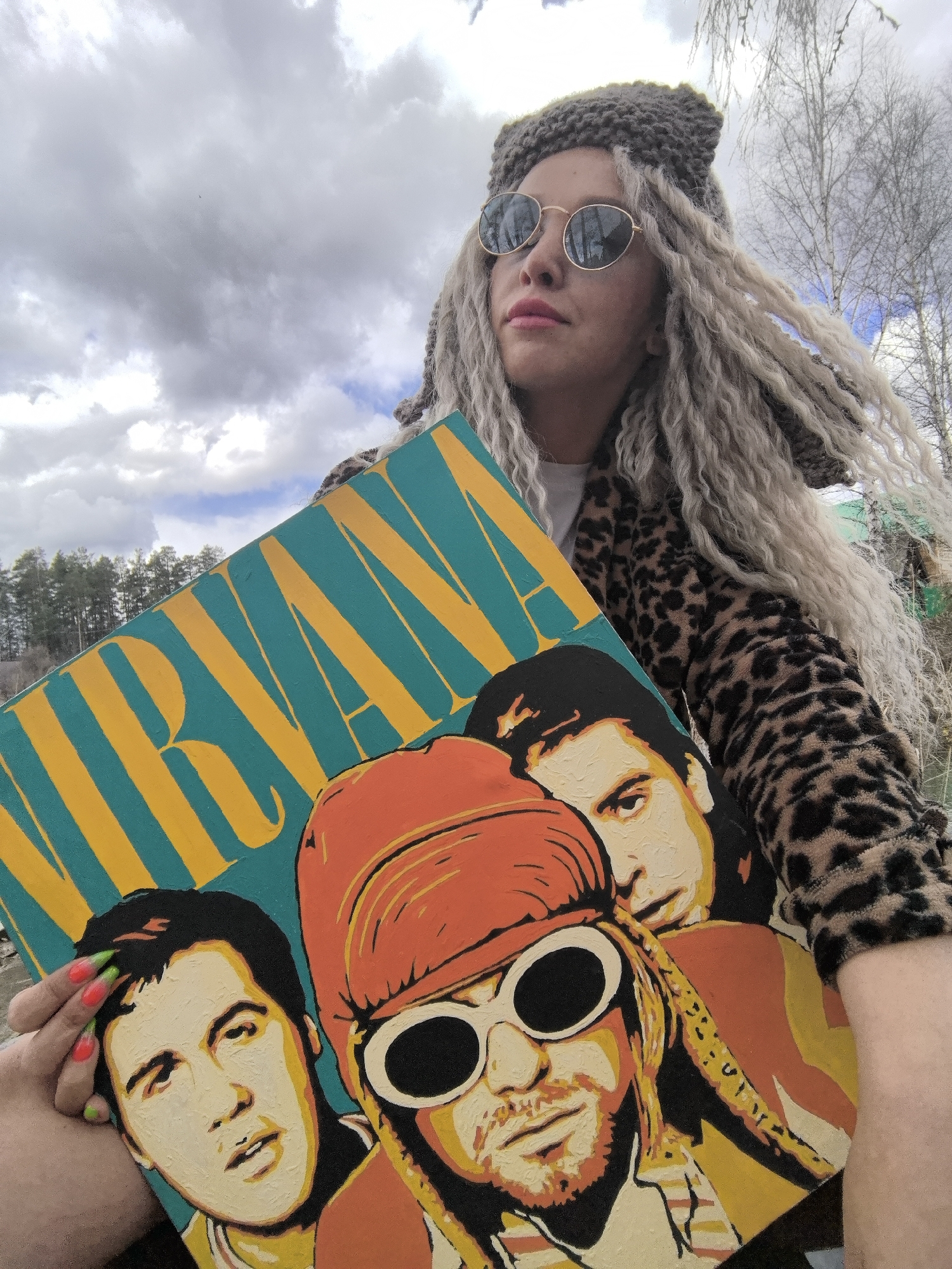 A person, likely a woman, is depicted outdoors, holding a vibrant, stylized painting of the band Nirvana. She has long, light blonde dreadlock-style hair, a chunky grey knitted beanie, round gold-rimmed sunglasses, and is wearing a leopard print jacket. Her gaze is directed to the right, with a calm or contemplative expression.
The artwork she holds features the word "NIRVANA" in large yellow capital letters against a teal background. Below the text, three stylized portraits of the band members are prominent: Kurt Cobain in the foreground with an orange beanie and white-rimmed sunglasses, flanked by Dave Grohl and Krist Novoselic. The painting utilizes bold colors and a distinct pop art aesthetic. The person's left hand, visible on the artwork, shows nails painted in bright orange with neon green tips.
The background consists of an overcast sky with some lighter blue patches, and a scattering of bare trees typical of late winter or early spring, with evergreen trees visible in the distant landscape. The setting is an outdoor, natural environment, with the location context being Noviy, Russia. The scene is illuminated by natural daylight.