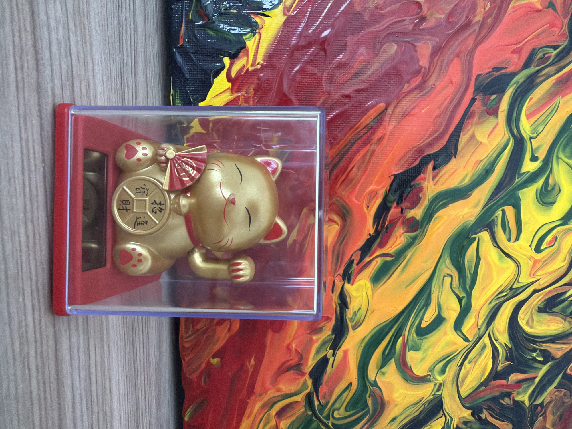 The image shows a golden "lucky cat" figurine in a clear plastic case, placed on a light-colored wooden surface. The cat has its eyes closed, a red bow around its neck, and is holding a coin with Chinese characters. Behind the figurine is a colorful abstract painting with swirls of red, yellow, black, and green. The painting appears to be textured, with visible brushstrokes and layers of paint. The setting suggests an indoor environment, possibly a display or a home. The overall mood is bright and cheerful, likely due to the colors of the painting and the auspicious nature of the lucky cat figurine. No specific time of day or weather conditions are discernible. The image is cropped from a larger view, with the wooden surface and the painting providing background context.