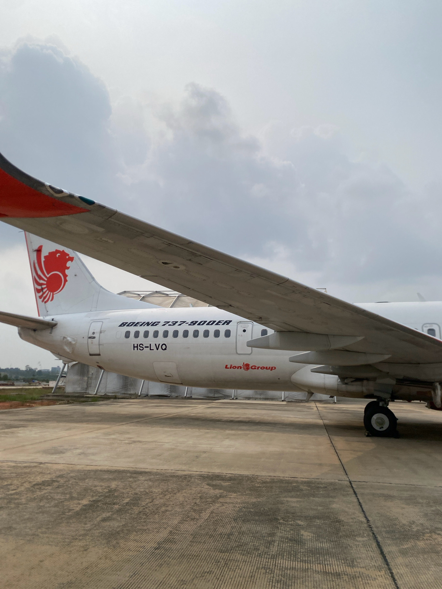 The image shows the rear section of a white Boeing 737-900ER aircraft with red and orange accents on the tail and wingtip. The aircraft is parked on a concrete tarmac. The Lion Group logo, a stylized red lion's head, is prominently displayed on the tail. The registration number "HS-LVQ" is visible on the fuselage, as well as the text "BOEING 737-900ER" and "Lion Group". In the background, there are white tent-like structures, possibly hangars or temporary shelters, and some vegetation. The sky is overcast with large, diffused clouds. The image appears to have been taken at Hang Nadim International Airport (BTH) in Batam, Indonesia, based on the presence of aircraft and airport infrastructure.