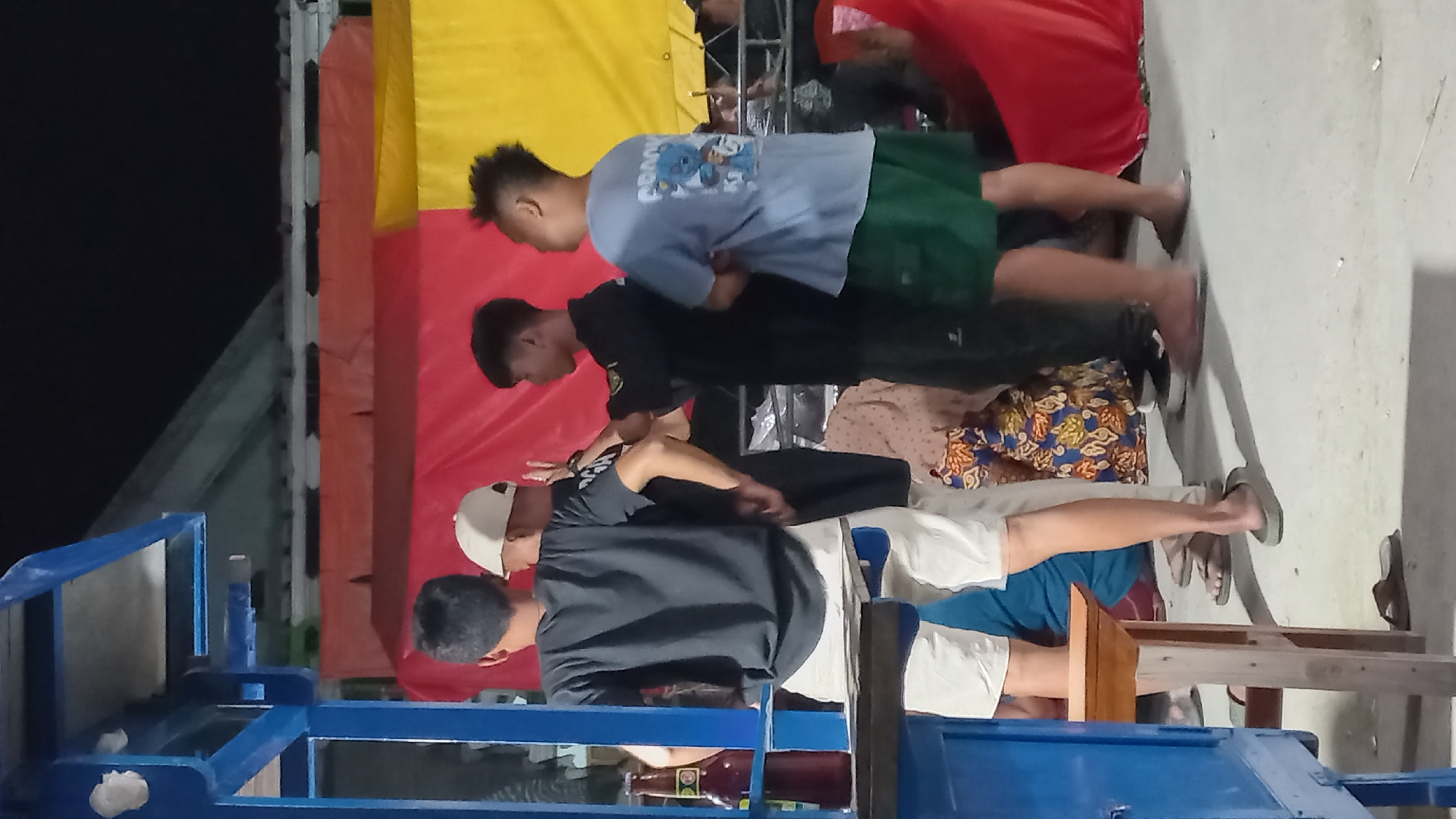 In Kedungmegarih, Indonesia, a group of four young men are gathered near a blue wooden structure. The man on the far left wears a dark t-shirt and white shorts, standing with his back to the camera. Next to him, a man in a white cap faces away from the viewer, with his arms raised slightly. The third man, wearing a black shirt and dark pants, also has his back to the camera, with his left arm extended. The man on the far right, in a light blue t-shirt and green shorts, is seen from the side, looking towards the center. Behind them, red and yellow tarpaulins are visible, suggesting an outdoor or semi-outdoor setting. A bottle of what appears to be a dark beverage sits on a ledge of the blue structure. The ground is concrete, and some patterned fabric is visible on the ground to the right. The scene is dimly lit, indicating it might be evening or night.