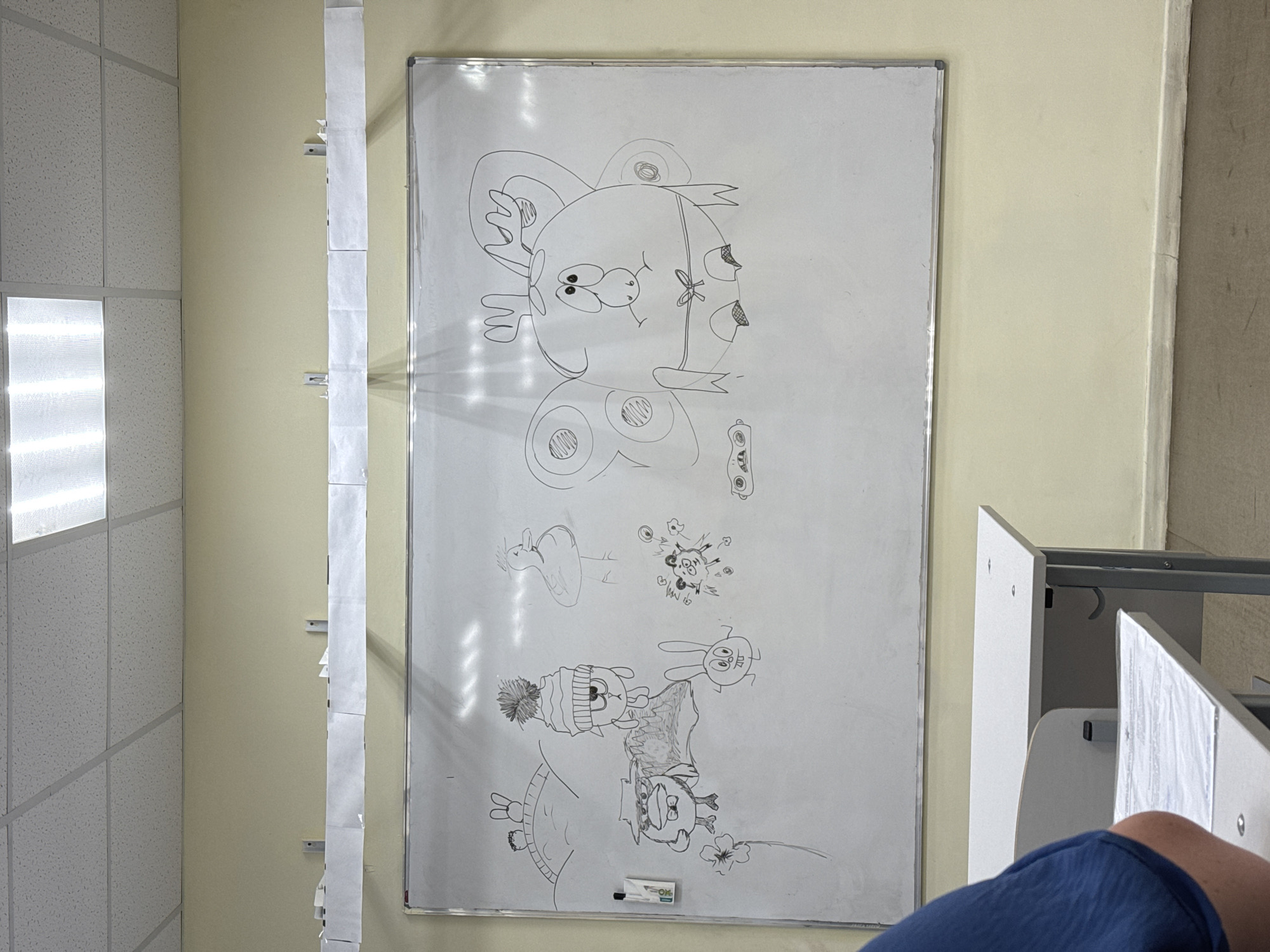 The image shows a whiteboard with drawings on it, mounted on a light yellow wall. To the left of the whiteboard is a wall with a tiled pattern, and on the right, there are desks and chairs. The drawings on the whiteboard include cartoon-like characters and flowers. A long strip of paper is attached to the wall next to the whiteboard. The location is Zav'yalovo, Russia. A person is partially visible in the lower right corner of the image.