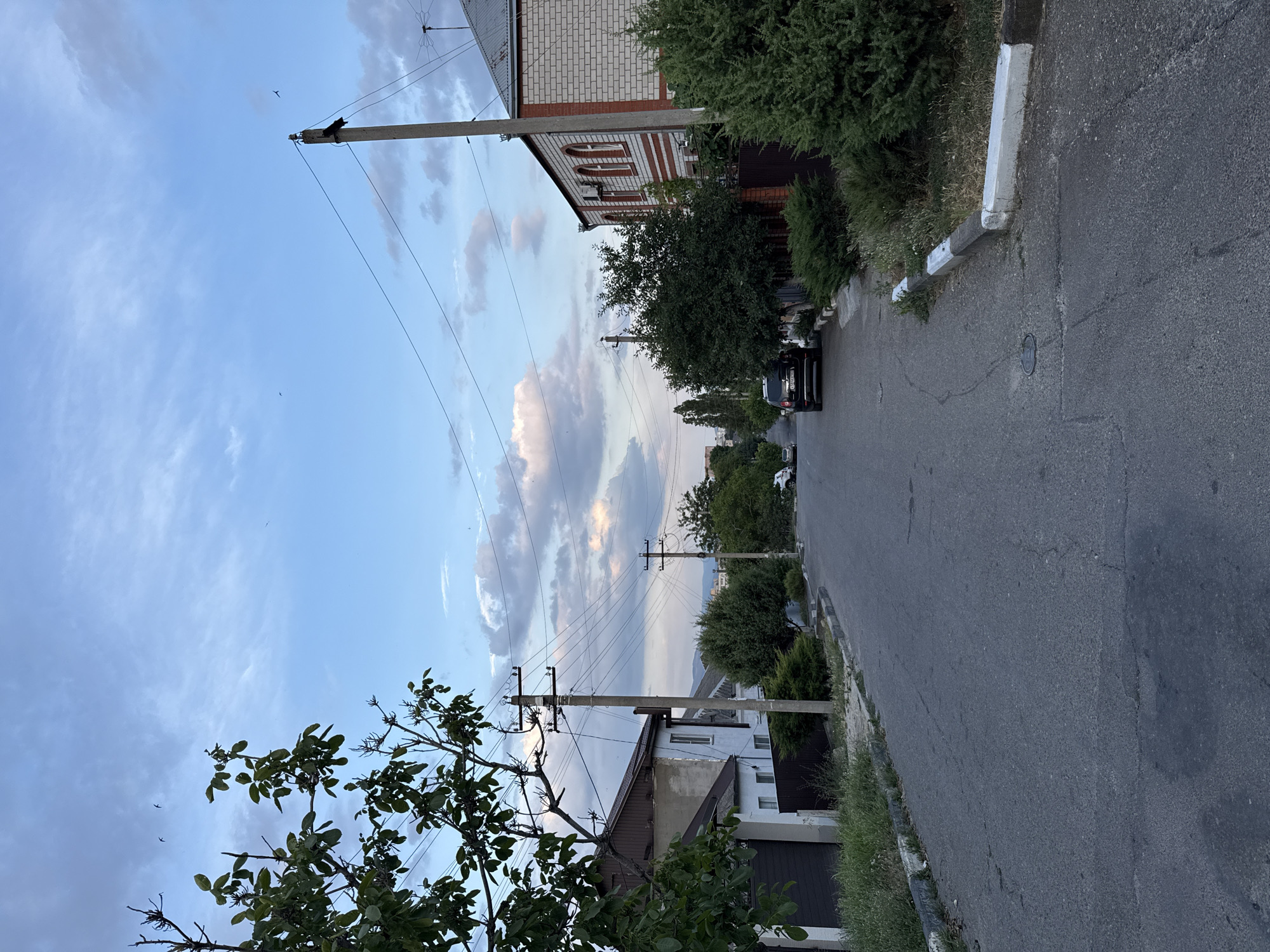 The image shows a street scene in Novorossiysk, Russia, under a partly cloudy sky. A paved road curves gently uphill, bordered by residential buildings and lush greenery. On the left, a house with a light-colored facade and a brown roof is visible, partially obscured by a tree with dark green leaves. Further down the street on the left, a fence and more vegetation line the road.
On the right side of the street, a two-story building constructed of white brick with red trim and arched windows stands prominently. A dark-colored SUV is parked on the right side of the road, facing uphill. Numerous utility poles with power lines crisscross the sky. The overall impression is a quiet, suburban street with a hint of natural beauty, bathed in the soft light of late afternoon or early evening.