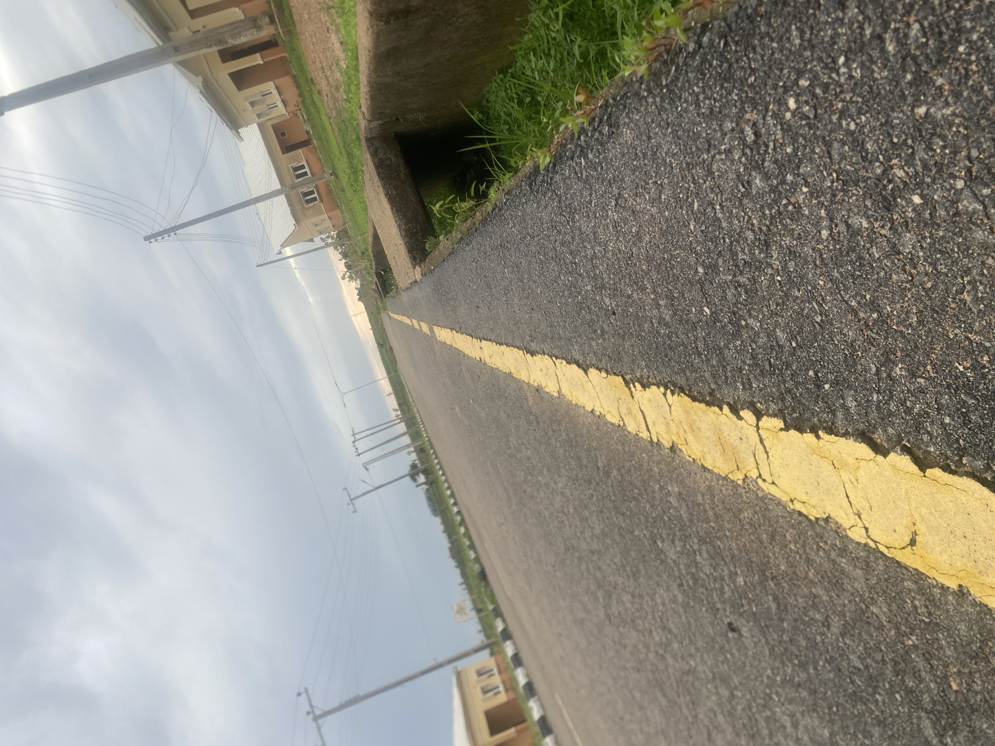 This image depicts an asphalt road with a yellow line marking its edge, stretching into the distance. The road is bordered by a concrete drainage channel on the left, filled with green grass and vegetation. Beyond the channel, several residential buildings with light-colored walls and brown accents are visible. Utility poles with power lines are also scattered along the left side of the road, suggesting an urban or suburban setting.
The perspective is low, angled towards the ground and the horizon, giving a sense of looking up from street level. The sky is overcast and appears to be either early morning or late afternoon, judging by the diffused light. The overall mood is somewhat subdued due to the cloudy sky.
The location is identified as Bulamare, Nigeria. The presence of residential buildings, paved roads, and utility infrastructure suggests a developed area within Nigeria. No people are visible in the image, and no specific activity or event is clearly taking place; it appears to be a static scene of a road and its surroundings. The image is rich in detail regarding textures and surfaces, with the rough asphalt, the smooth concrete, and the organic textures of the grass all being distinctly visible.