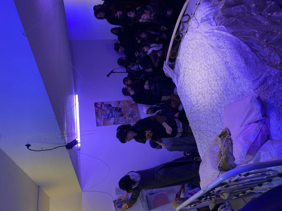 A crowded room with a blue light illuminating a group of people standing near a white metal bed with a patterned cover and pillows. Some individuals are wearing VR headsets. Paintings adorn the wall behind them, and a microphone is visible. A fluorescent light fixture is attached to the ceiling, casting a blue glow over the scene.