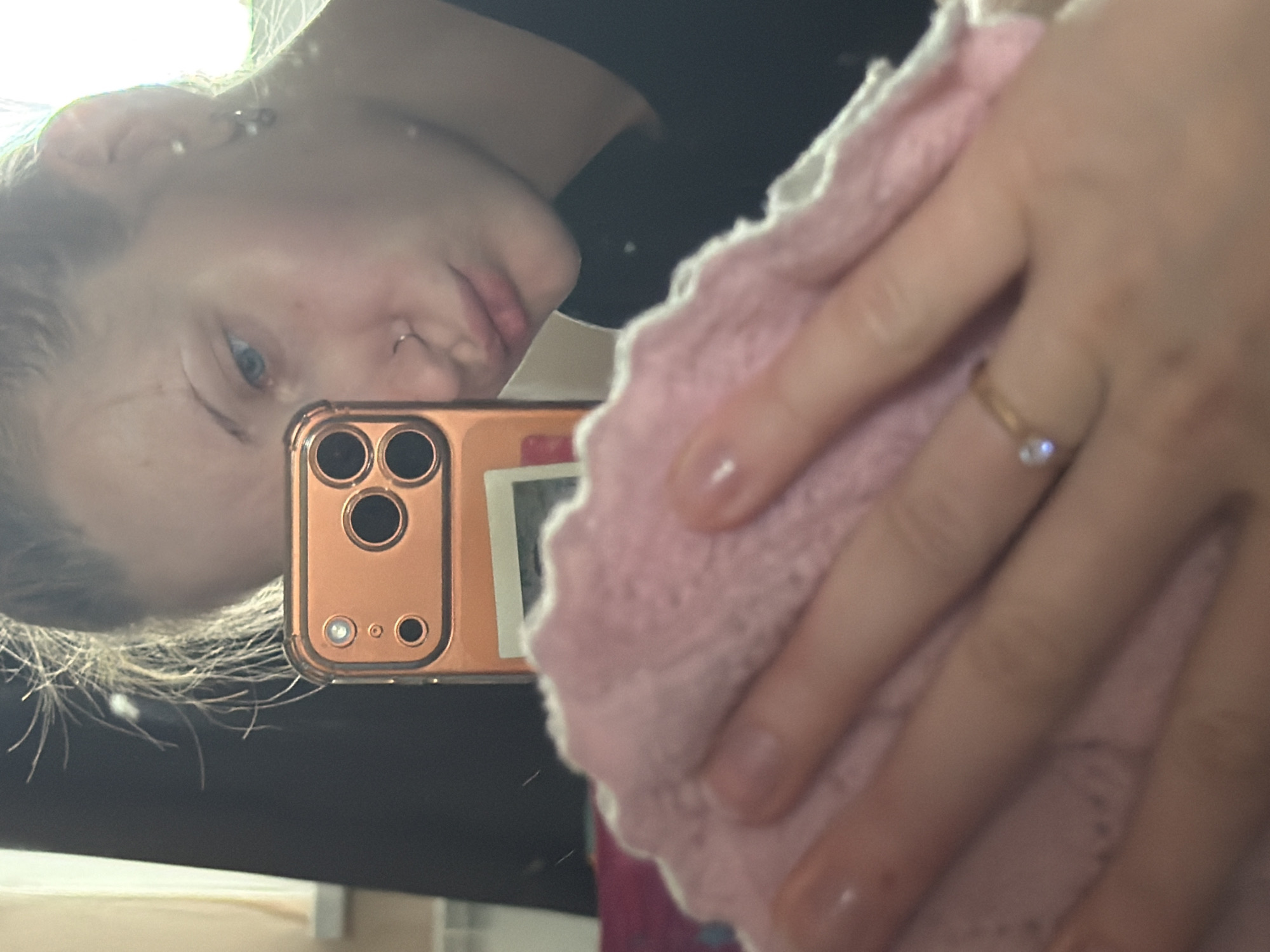 The image is a close-up, slightly over-exposed, vertical, mirror selfie. The primary subject is a person's face, reflected in a mirror. They have fair skin, blue eyes, and a nose ring. Their hair appears blonde and is partially visible around the edges of the frame. The person is holding a pink, knitted material in their hands, which obscures part of their face and the mirror's surface. On their left ring finger, they are wearing a thin gold ring with a small, clear stone.
In the reflection, a cell phone is visible, held by the person taking the photo. It appears to be an iPhone with an orange or rose gold casing, featuring a multi-lens camera system. A small, white square object, possibly a sticker or a small photo print, is affixed to the back of the phone case. The overall lighting suggests it might be daytime, possibly indoors due to the reflective surface. The mood appears casual and personal. No visible text is present in the image.