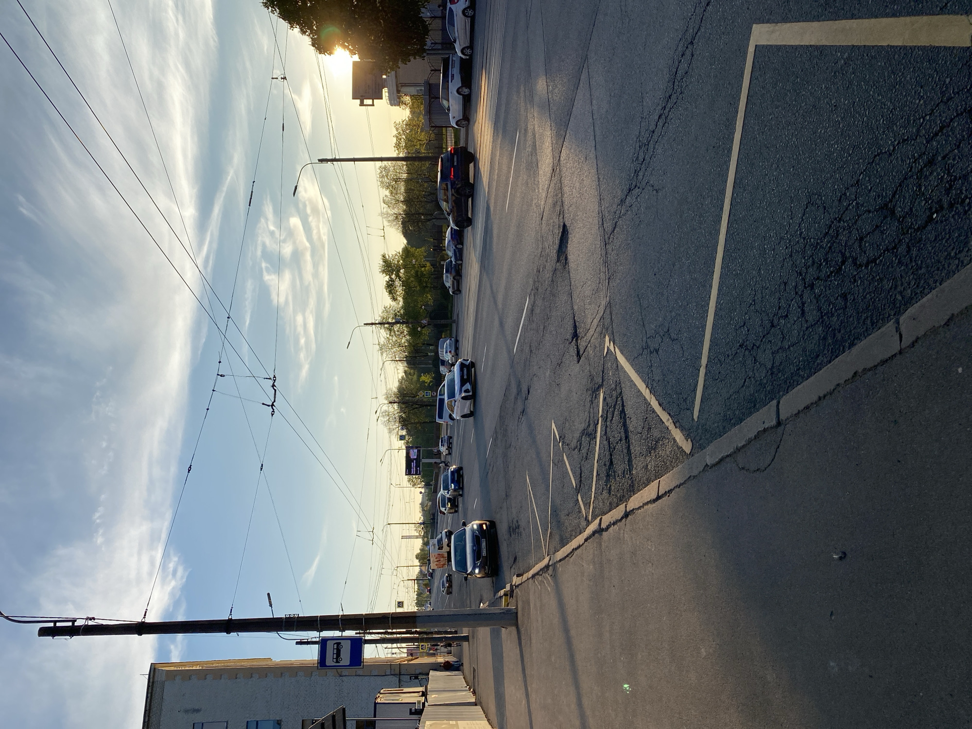 The image depicts a street scene in Saint Petersburg, Russia, at what appears to be late afternoon or early evening based on the lighting. The view is looking along a road with multiple lanes of traffic. Cars are lining up, likely due to traffic congestion. The road surface is cracked and shows signs of wear. There are tram or trolleybus overhead wires visible, which suggests the presence of public transportation. There are light poles on the side of the road. A bus stop sign is visible in the lower left corner. There are buildings on either side of the road. The sky is clear with some wispy clouds and the sun is shining through, partially obscured by buildings.