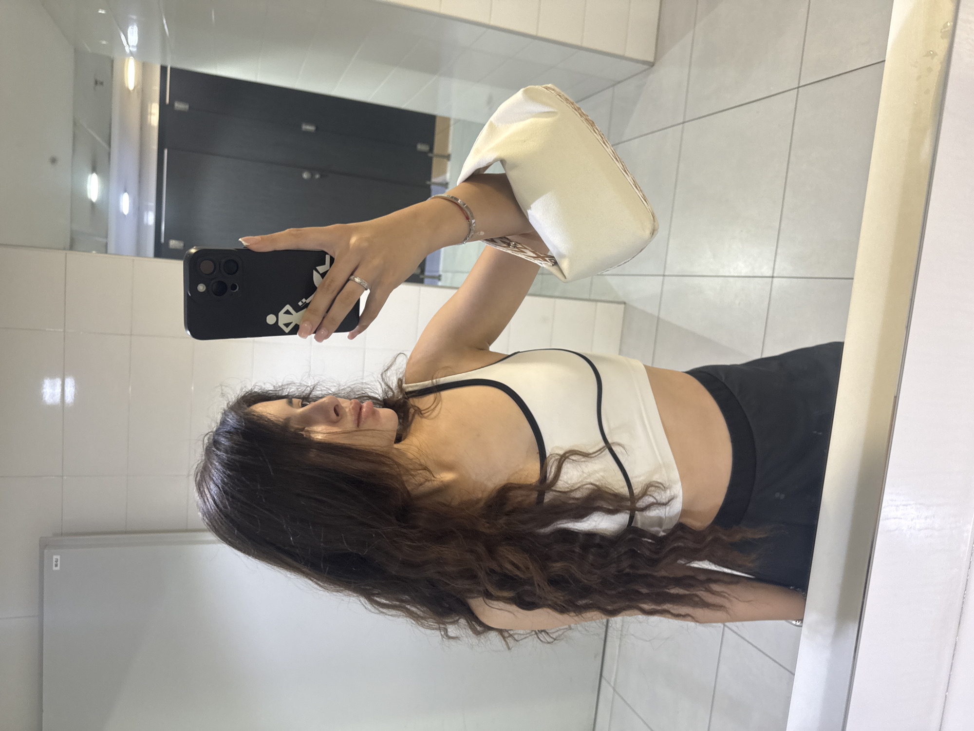 A young woman with long, dark, curly hair stands in a brightly lit, tiled bathroom, taking a mirror selfie. She is wearing a white sports bra with black trim and black shorts. She holds a black smartphone in her right hand, angled to capture her reflection and a small, cream-colored handbag. A silver bracelet adorns her left wrist, and a silver ring is on her right ring finger. The bathroom environment features white tiled walls and floor, with dark stall doors visible in the background. The scene suggests a private or public restroom facility, possibly in a gym or a similar establishment, located within the city of Plovdiv, Bulgaria.