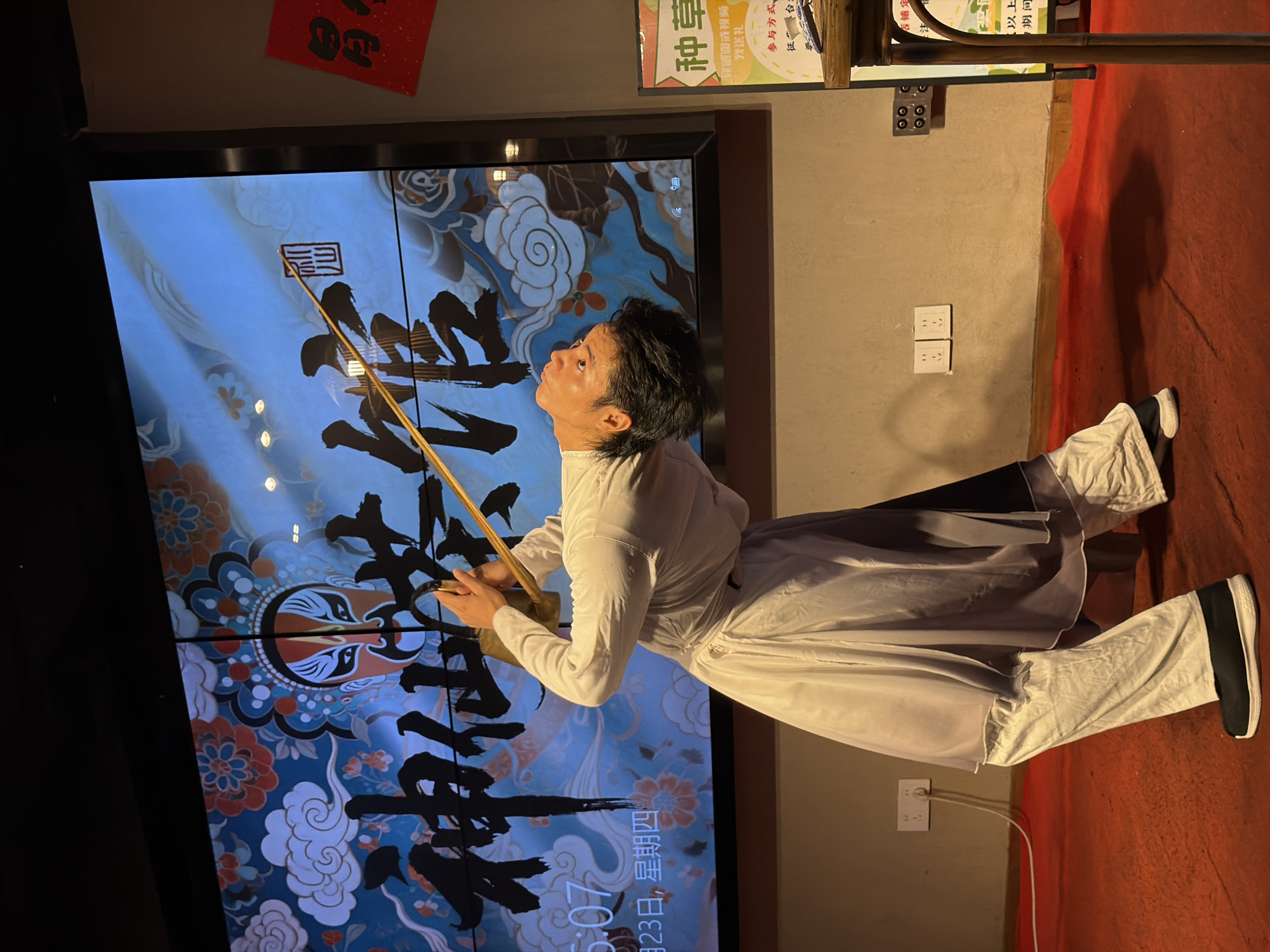 A male performer, dressed in traditional white and gray layered attire, is shown mid-performance, bending backward with a long wooden pole balanced vertically on his head and chin. The backdrop is a large screen displaying a vibrant, colorful image featuring a Peking Opera mask and Chinese calligraphy. To the right of the screen, a red sign with Chinese characters is visible, and a yellow promotional poster with text and illustrations is also present. The performer is standing on a red carpeted floor, with electrical outlets and cords visible on the wall behind him. The overall scene suggests an indoor cultural performance or demonstration in Yuzhong District, China.