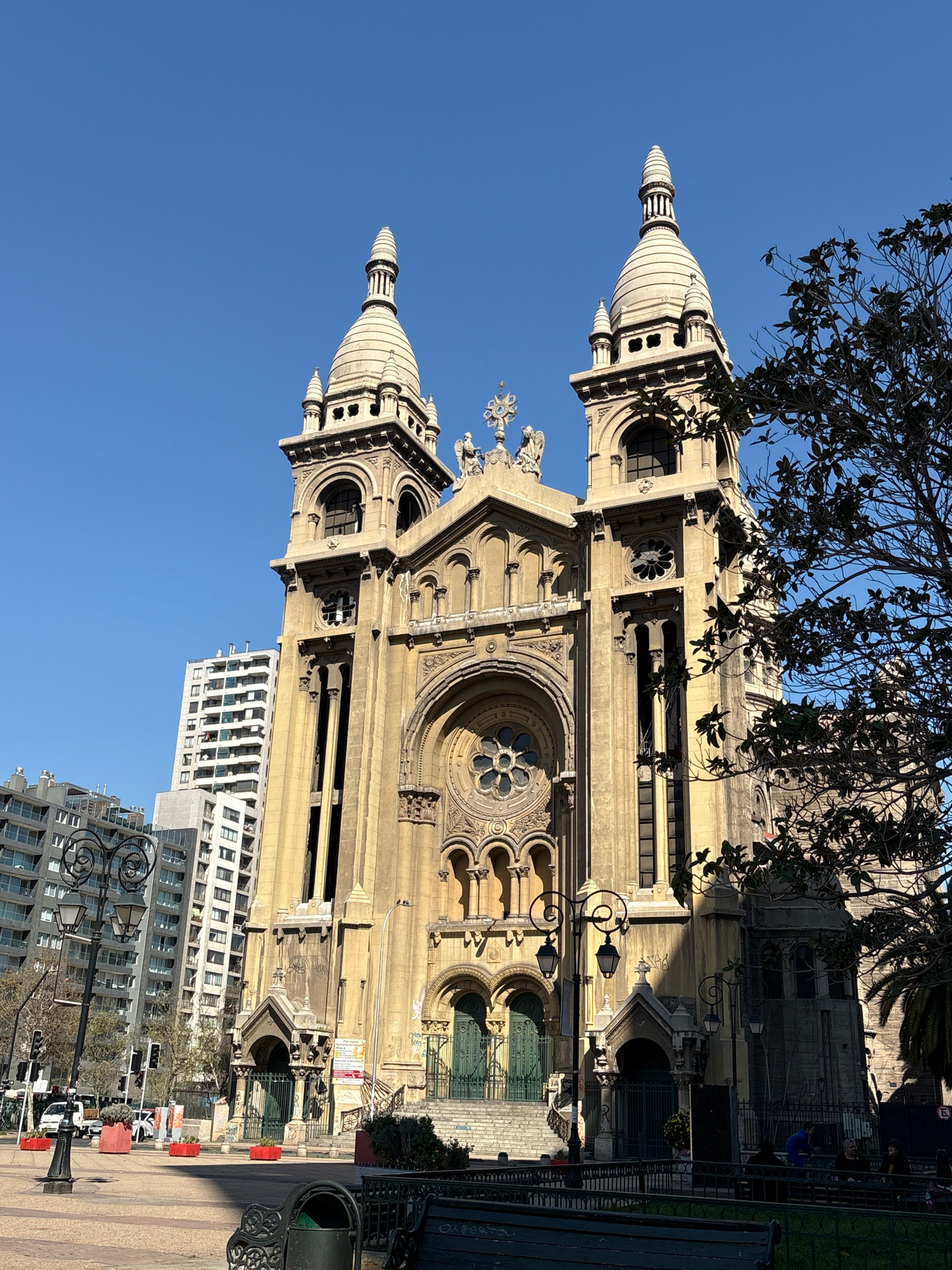 The image shows the Metropolitan Cathedral of Santiago, Chile, a prominent religious building with a grand facade and twin towers topped with spires. The cathedral is made of stone, with intricate architectural details. In front of the cathedral, there is a public plaza with lampposts, benches, and planters. Modern high-rise buildings are visible in the background, suggesting an urban setting. The sky is clear and blue, indicating a sunny day. There are a few people in the plaza, some sitting on benches and others walking. The overall scene depicts a sunny day in a bustling urban environment, with the historic cathedral serving as the focal point.
