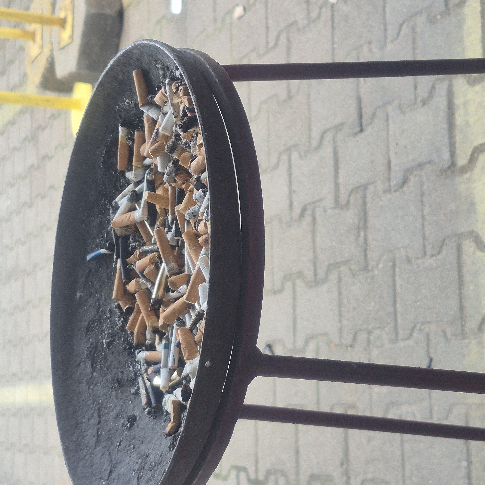 An ashtray overflowing with cigarette butts sits on a stand in front of paving stones and yellow bollards. The ashtray is made of black metal and is a little over half full with cigarette butts, some of which have been extinguished and are covered in ash. The butts are a mix of filtered and unfiltered, with some bearing colored bands. One blue cigarette filter is visible standing upright in the center of the ashtray. The stand supporting the ashtray is made of dark metal and has three legs. The paving stones are a light gray interlocking pattern. Two yellow bollards are visible in the background, one slightly out of focus behind the ashtray. The scene appears to be outdoors, possibly in a public area.