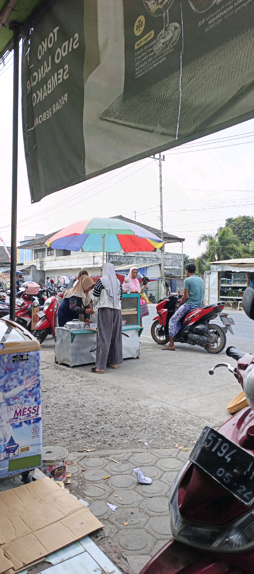 The image depicts a street scene in Kasiyan, Indonesia, with a focus on a food stall. In the foreground, a red motorcycle is partially visible, its license plate reading "5194 - II 05 - 24". To the left, a blue vending machine with an advertisement featuring "LIONEL MESSI" is present, along with a cardboard box. A gray trash bin with some debris around it is also visible. The main action centers on a food stall with a colorful umbrella overhead. Two women are attending the stall; one, wearing a hijab and striped shirt, is interacting with a customer. Another woman, wearing a pink hijab, stands behind the stall. A customer, wearing a blue t-shirt and patterned pants, is seated on a red motorcycle to the right, facing the stall. In the background, several other motorcycles are parked, and buildings line the street. The ground is paved with hexagonal tiles and asphalt. The sky is overcast.