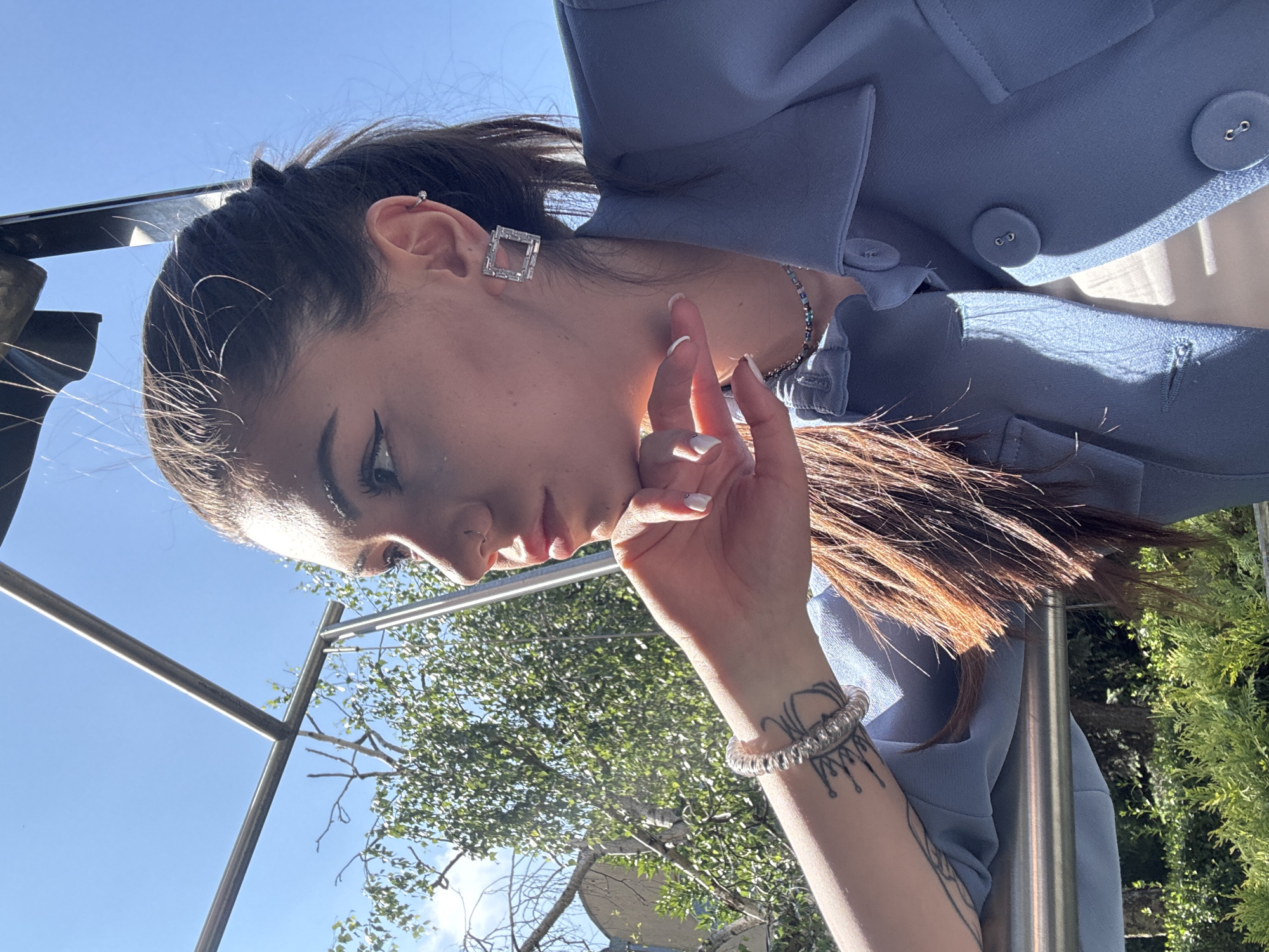 An outdoor portrait captures a young woman from Sofia, Bulgaria, in a rotated, almost horizontal orientation. She has dark hair styled in a high ponytail and wears a light blue blazer over a white top. Her accessories include a prominent square, gem-studded earring in her left earlobe, a small stud in her upper ear, a thin silver necklace, and multiple bracelets on her left wrist, one beaded and another coiled and clear. A distinctive dark, stylized tattoo is visible on her inner left forearm. Her fingernails are painted white.
The woman is shown in profile, looking downwards with a thoughtful or contemplative expression. Her left hand is raised, with fingers lightly touching her face near the chin, which features visible makeup, including eyeliner and glitter on her cheekbone.
The background consists of a clear, bright blue sky dominating the upper portion of the frame, and vibrant green foliage and trees in the lower section, indicating an outdoor, sunny setting. A sturdy metal railing or structure runs through the scene. The bright daylight casts strong highlights and shadows, accentuating details on her clothing and face. The scene appears to be an urban outdoor space, possibly a park or balcony. No visible text is present in the image.