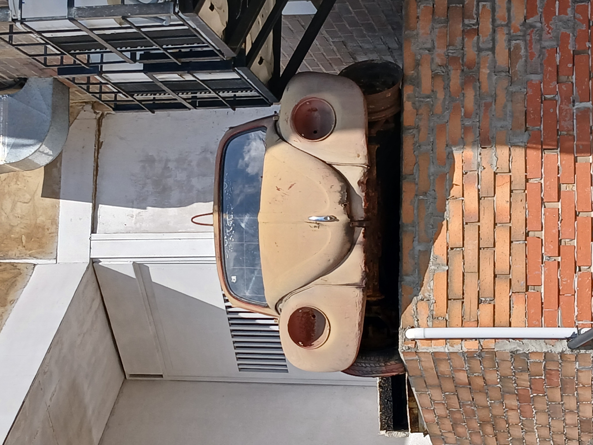 The image displays a weathered, beige Volkswagen Beetle (Type 1) parked outdoors, with its front facing the viewer. The classic car exhibits significant rust and faded paint, and its circular headlights are notably absent. To the right of the vehicle, a reddish-brown brick wall stands, featuring a white pipe horizontally attached. Portions of the wall are shaded, indicating bright daylight, likely midday or early afternoon, under a clear sky.
On the left side of the scene, a large, curved metal duct and a section of a white building wall are visible. Above the car, a black metal grate structure spans the upper part of the frame, possibly part of an overhead walkway or industrial installation. A rusty metal barrel is partially visible behind the car, adjacent to the brick wall.
The overall setting suggests an older, possibly industrial or neglected urban environment in Venezuela. The car appears stationary and disused. No people are present, and no discernible text is visible anywhere in the frame.