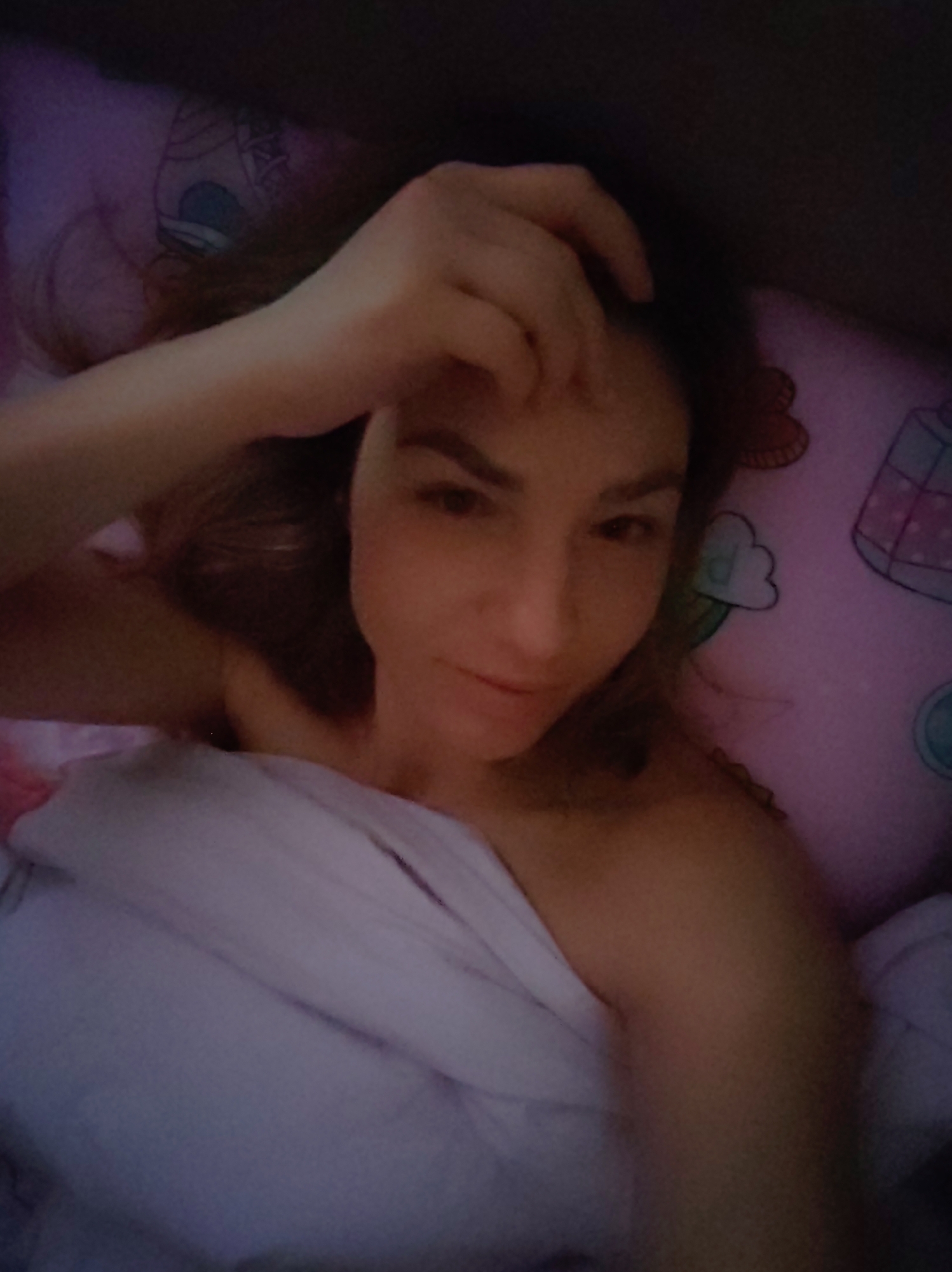The image shows a woman lying in bed, capturing a close-up, top-down perspective, likely taken as a selfie. She appears to be relaxing or possibly waking up, with a soft, slightly smiling expression. Her left arm is raised, with her hand resting gently on her forehead or tangled in her dark hair. She is partially covered by a light-colored, possibly white or very light purple, blanket or sheet, revealing her bare shoulder.
The setting is a bedroom, specifically focusing on the bed. A pink pillow with various cartoon-like illustrations, including what appears to be cupcakes, clouds, a heart, and a small building or house, is visible beneath her head and to her right. The lighting is dim, suggesting it might be early morning, late evening, or night. The overall color palette is muted, with shades of purple, pink, and dark tones. There is no visible text in the image itself. The context provided indicates the location is Saint Petersburg, Russia.