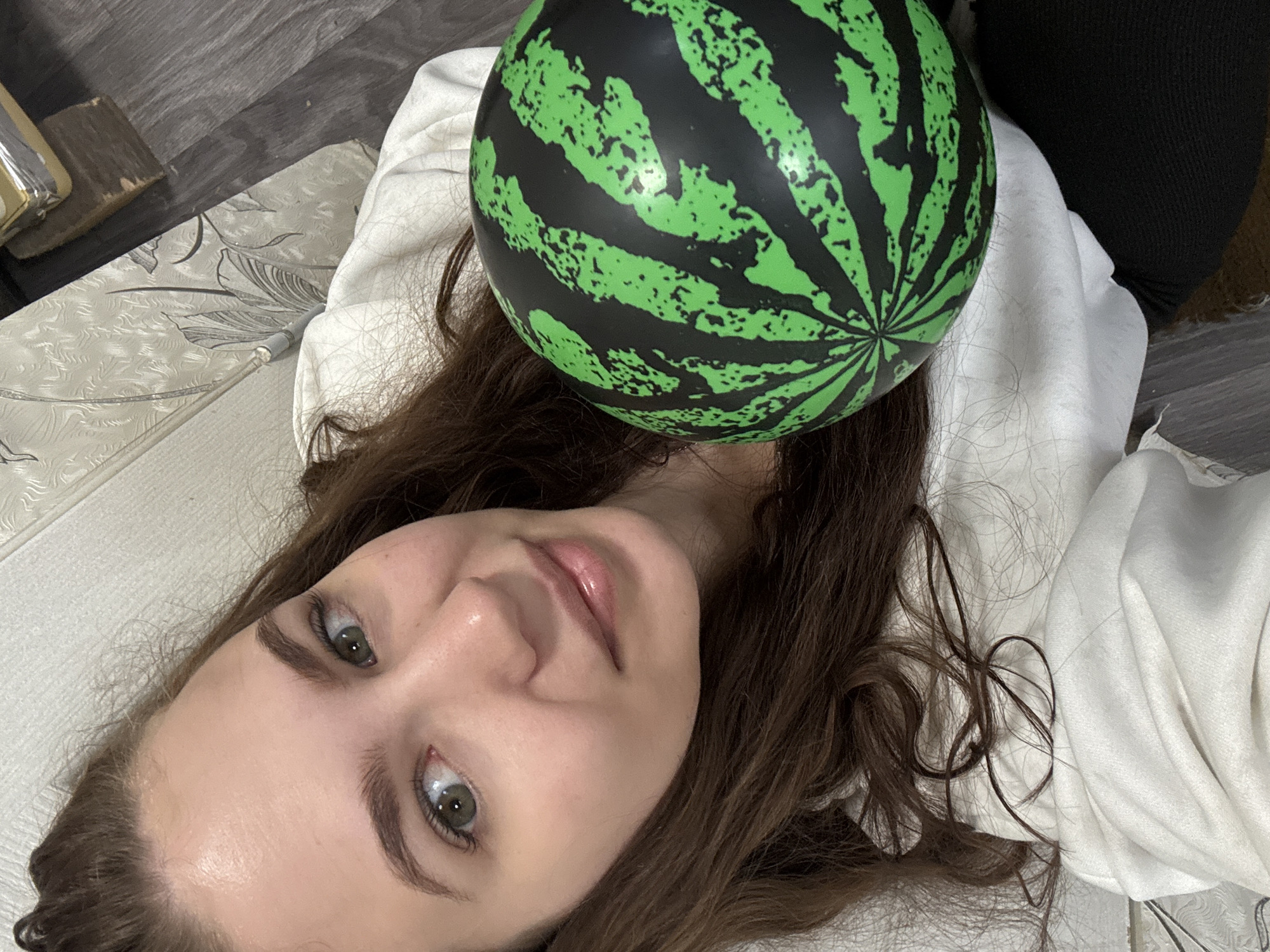 A young, fair-skinned woman with brown hair and green eyes lies on a patterned surface in an indoor setting. She is looking directly at the camera with a slight smile. A green and black watermelon-patterned ball rests on her head. She is wearing a white garment. The background includes a wooden floor and some items in a corner. There is no verifiable evidence to definitively confirm that the image was taken in Nizhnyaya Gar', Russia.