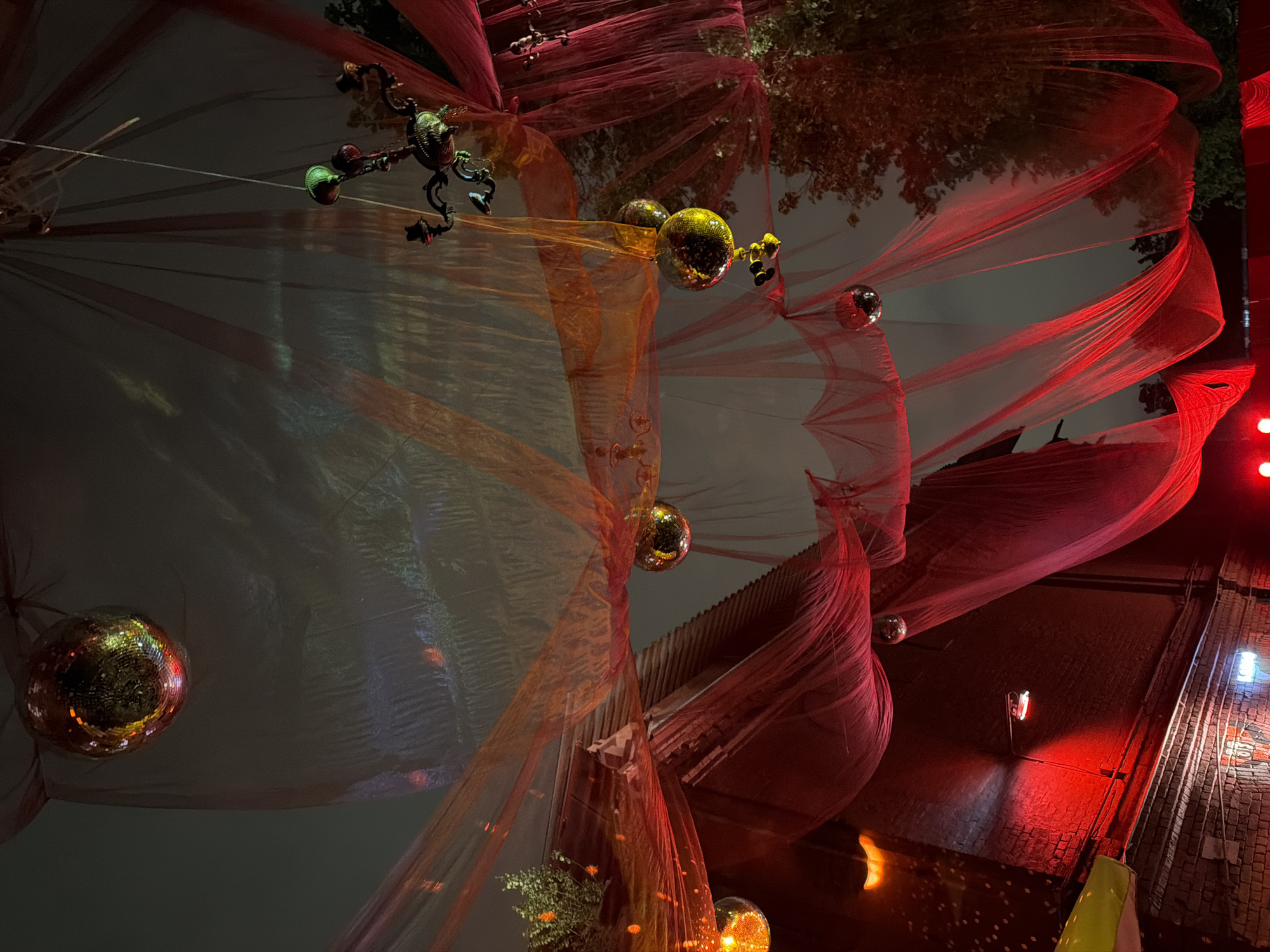 The image depicts a street scene at night, possibly in Moscow, Russia. It features a red, translucent fabric draping overhead, adorned with disco balls. An ornate metal fixture with green bulbs hangs from the fabric. In the background, there are trees and buildings. The overall atmosphere is festive and illuminated with red lighting. A brick building is visible on the right.