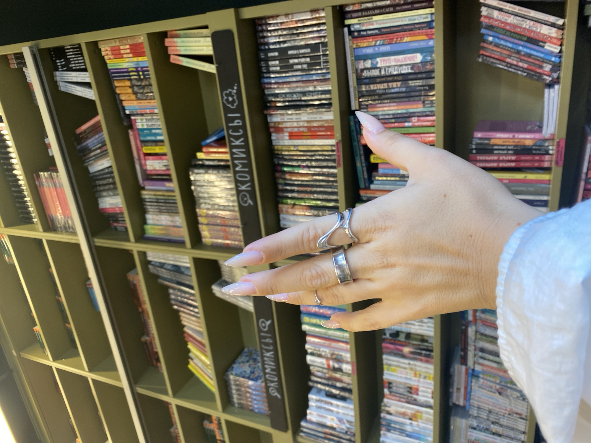 A close-up shot focuses on a person's left hand adorned with several silver rings, positioned in front of a library shelf filled with books. The hand is positioned in the foreground, with the fingers slightly curled and extended, showing long, manicured nails. The rings are distinct: a thin band on the index finger, a wider band on the middle finger, and a uniquely shaped, possibly abstract design on the ring finger. The shelves, painted a pale green, are densely packed with books of various sizes and colors, suggesting a bookstore or a public library. Some books are oriented vertically, others horizontally, creating a textured backdrop. A dark sign with white lettering that reads "КОМИКСЫ" (Comics) is visible above a row of books. The lighting suggests an indoor environment, possibly with ambient light from the side, casting soft shadows. The image provides no clear indication of the specific location within Saint Petersburg, Russia.