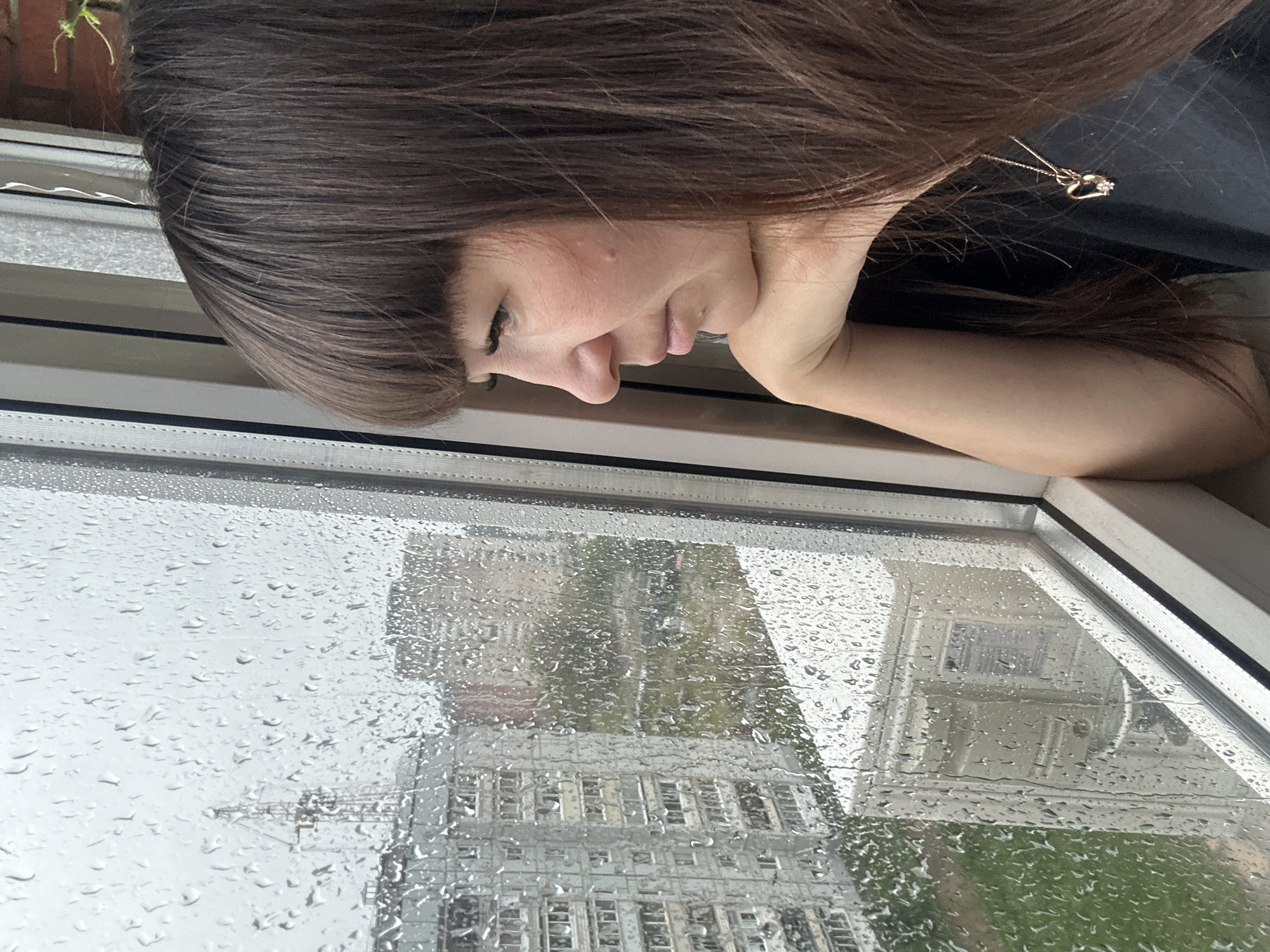 A woman is looking out of a window at apartment buildings in Chaikovsky, Russia. The window is wet with raindrops, obscuring the view of the city. The woman has long, dark brown hair and is wearing a dark shirt with a heart-shaped pendant. Her arm is resting on her chin as she gazes out at the rain. The apartment buildings are typical of Soviet-era architecture, with uniform facades and balconies. The sky is overcast, and the overall mood of the image is somber.