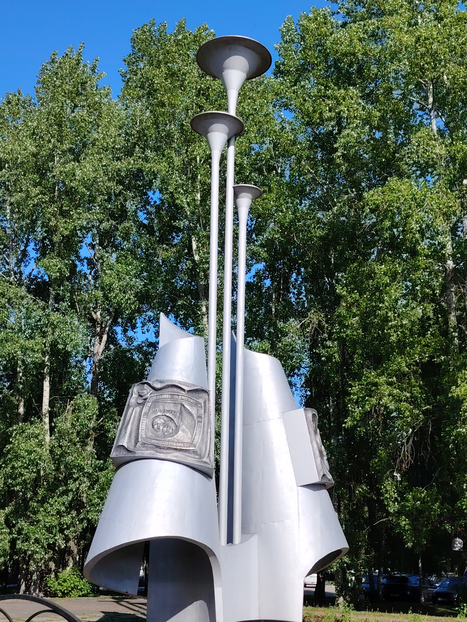 The image shows the "Memorial Sign" in Chaikovsky, Russia. It is a large metal sculpture consisting of three tall, slender, lamp-like structures with concave shades at the top. These structures are interspersed between abstract geometric shapes that hold detailed, bas-relief carvings, possibly depicting historical or cultural themes. The sculpture is positioned outdoors with a backdrop of lush green trees and a clear blue sky. The foreground reveals a glimpse of the surrounding park or green space.