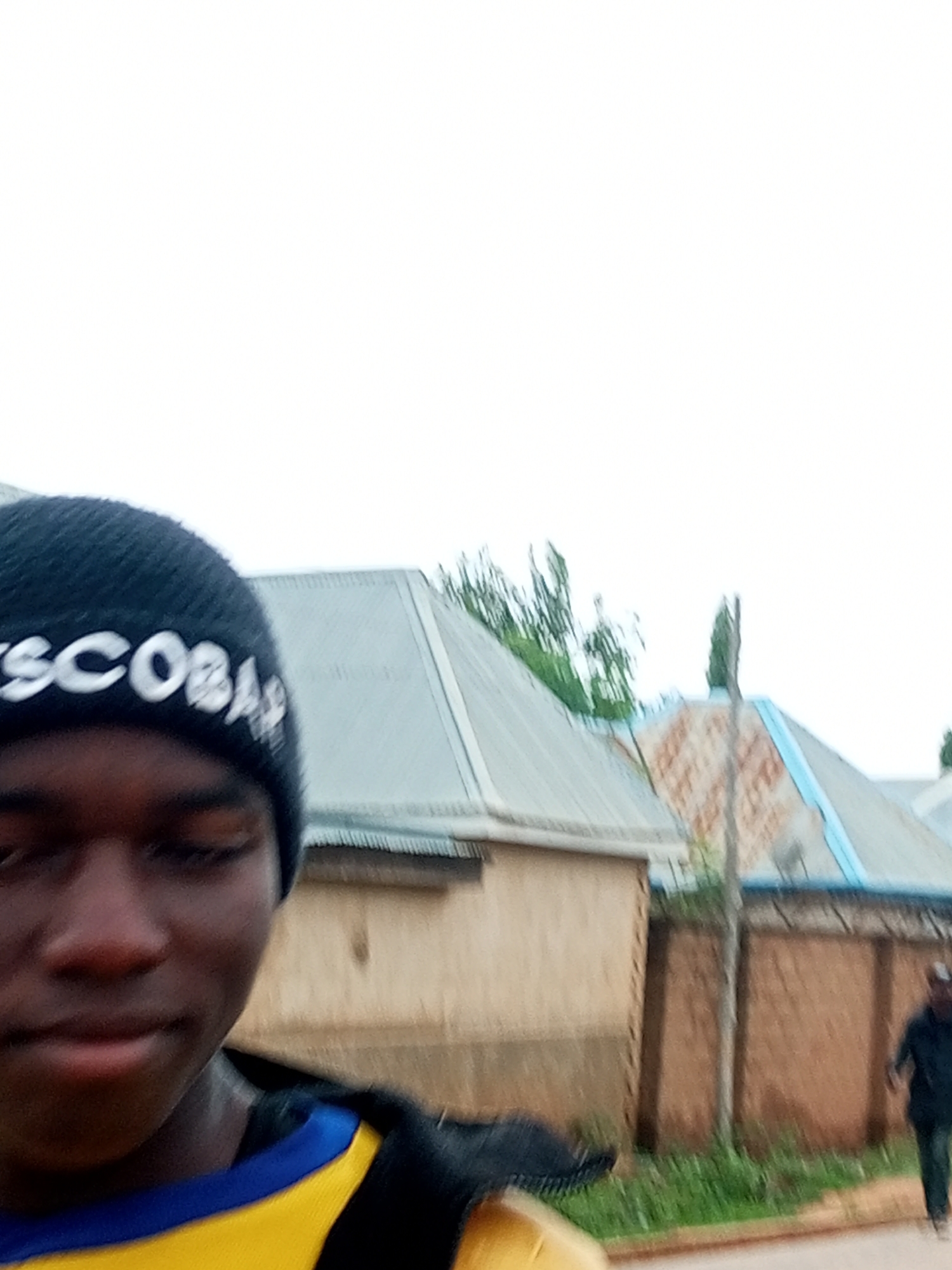 The image shows a close-up of a young person wearing a black beanie with white lettering that reads "SCOBAR". The person's face is partially visible, with their eyes closed and a slight smile. They are wearing a yellow and blue shirt. In the background, there are buildings with corrugated metal roofs, and some trees are visible. Further in the background, to the right, a person in a dark jacket and pants is walking away from the camera. The overall scene appears to be outdoors, possibly in a residential area. The location in the city of Tempure, Nigeria, is not explicitly identifiable from the visual elements alone.
