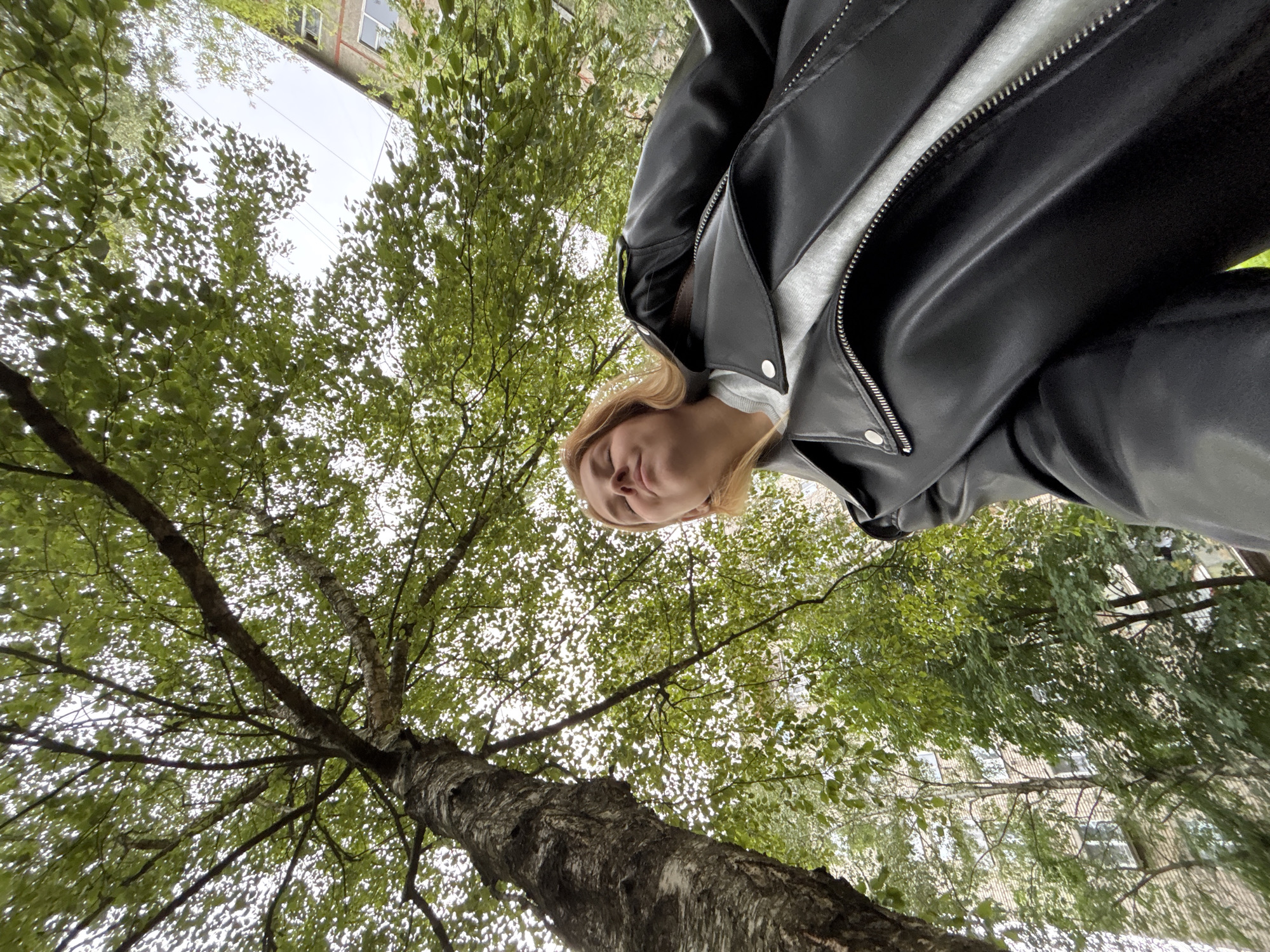An outdoor, low-angle, medium shot shows a young woman from the chest up, looking upward with her eyes closed and head tilted back. She has blonde hair and is wearing a light gray sweatshirt under a black leather jacket. The jacket is unzipped, exposing the collar of the sweatshirt. The background consists of the trunk and branches of a large tree with green leaves, and beyond that, a building and the sky. The scene is set in Rusanovo, Russia.