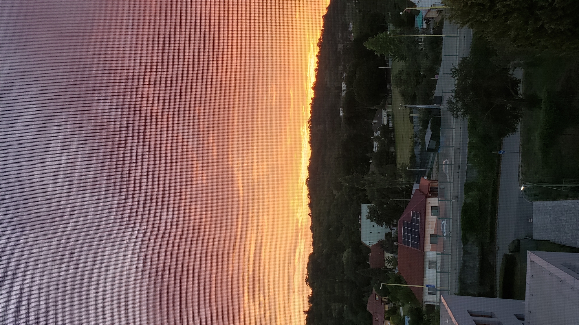 The image depicts a sunset over a residential area in Prešov, Slovakia. The sky is a gradient of purples, oranges, and yellows, with thin clouds stretched across the horizon. A dense line of trees forms the base of a hill, obscuring most of the natural landscape. In the mid-ground, several houses are visible, one with solar panels on its roof. A road runs horizontally across the scene, with a fence separating it from the houses. In the foreground, part of a modern building with a gray facade is visible. The overall impression is a calm, suburban evening scene.