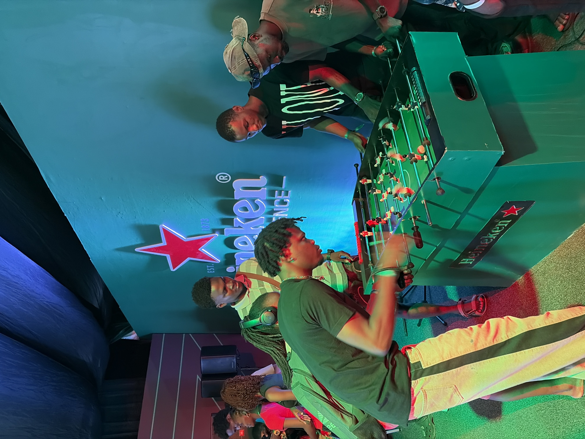 Several people are playing foosball in an indoor setting, possibly Lagos, Nigeria. The foosball table is green and features Heineken branding. There's a large backdrop with the Heineken logo. The players include individuals of African descent. Some are wearing casual clothing such as t-shirts and pants with stripes. There is a speaker visible in the background. Lighting appears to be from various sources, including some neon or colored lighting creating green and red hues. The overall impression is a recreational area sponsored by Heineken.