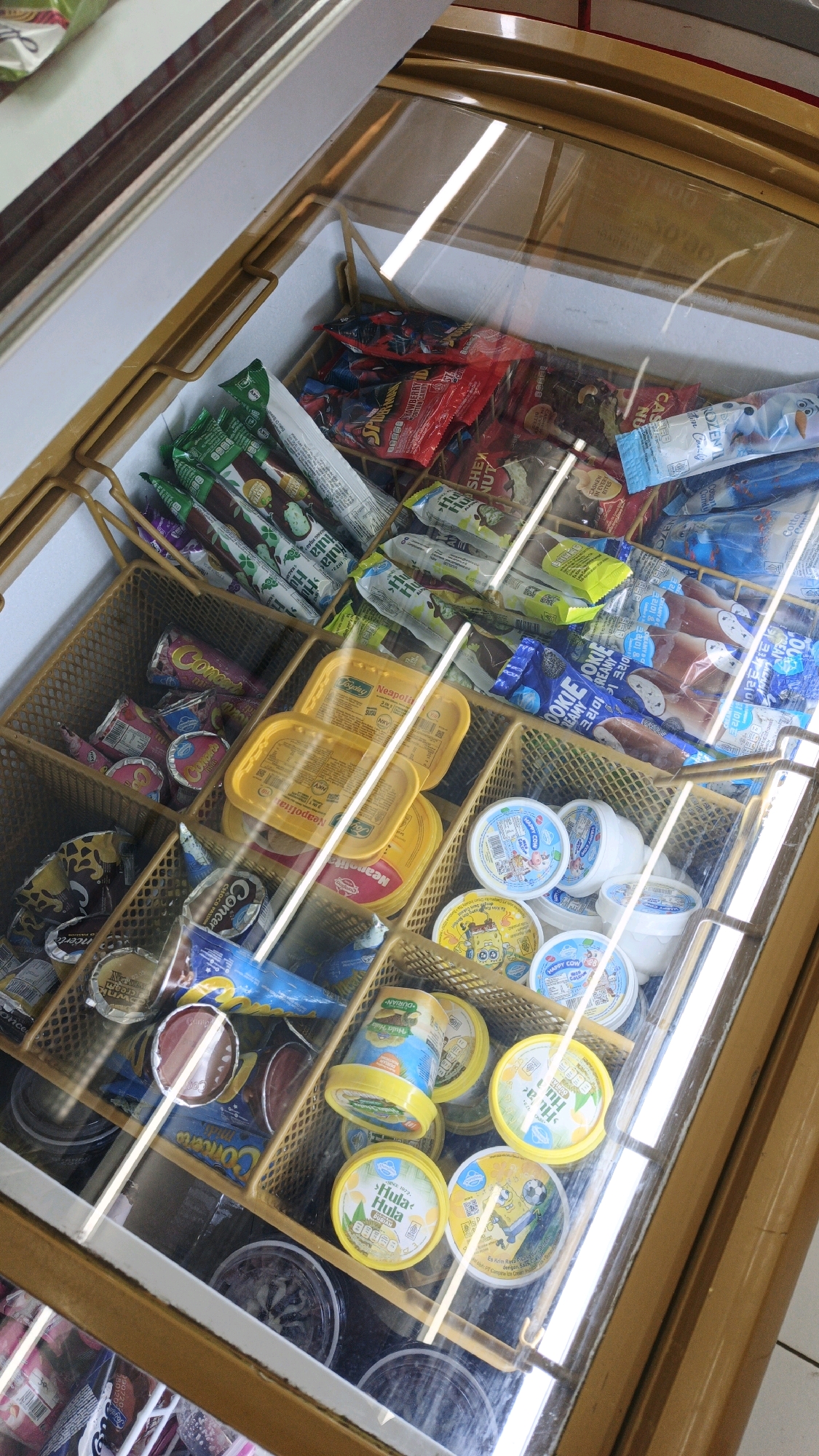 This is a high-angle, eye-level, indoor shot of a freezer chest filled with various ice cream products. The freezer is divided into compartments, each containing different types of frozen treats. In the foreground, there are several round tubs of ice cream with colorful labels, some featuring cartoon characters and the text "Hula Hula" and "Happy Cow." Behind these, there are rectangular yellow tubs, possibly butter or margarine, labeled "Neopolitana." The midground displays a variety of individually wrapped ice cream bars and cones. Some have colorful wrappers with cartoon imagery, including characters that appear to be from the Spider-Man franchise. Others are plain white or green, with some text visible in what appears to be Indonesian or Malaysian. In the background, more ice cream bars and cones are visible, some with chocolate coatings and others with colorful wrappers. The freezer has a glass lid, through which the contents are viewed. The overall environment appears to be a retail setting, possibly a convenience store or minimarket, as suggested by the organized display of products. The lighting is artificial, likely from the store's overhead lights and the freezer's internal lighting, creating reflections on the glass lid. The image does not contain any people or specific landmarks that would definitively place it in Parigi, Indonesia, though the packaging with Asian scripts hints at a Southeast Asian location.