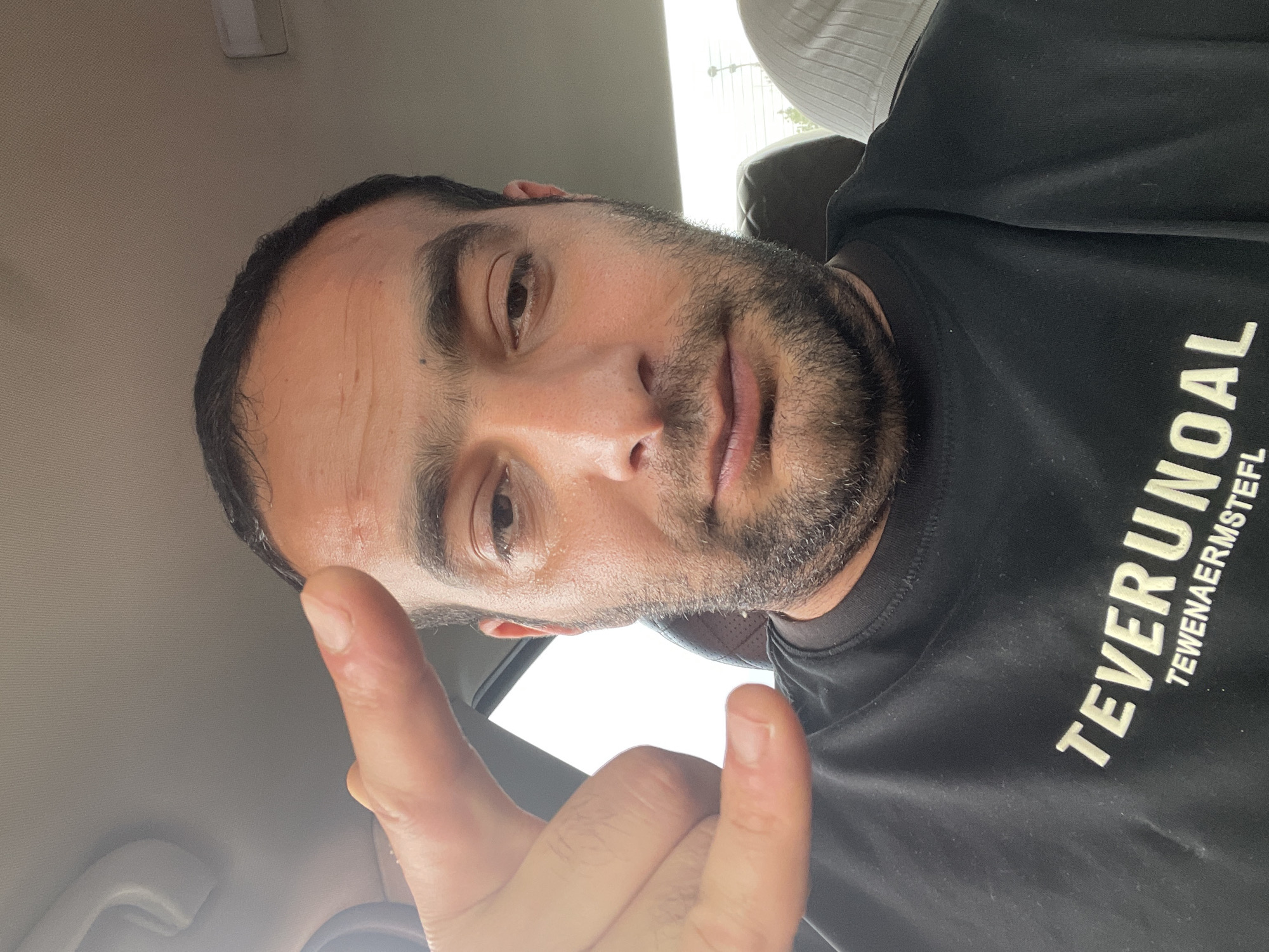 This is a selfie taken by a man inside a car. He is the sole subject. The man has dark hair, a beard, and is wearing a black t-shirt with white text on it. The text on his shirt appears to be "TEVERUNOAL" and "TEWENAERMSTEFL". The man is looking at the camera and his left hand is raised in the frame, with his index and pinky fingers extended in a "rock on" gesture. The background is blurry but suggests the interior of a car, with a light-colored headliner and a hint of sunlight coming through the windows. The lighting suggests it is daytime. The image is taken from a low angle, looking up at the man's face.