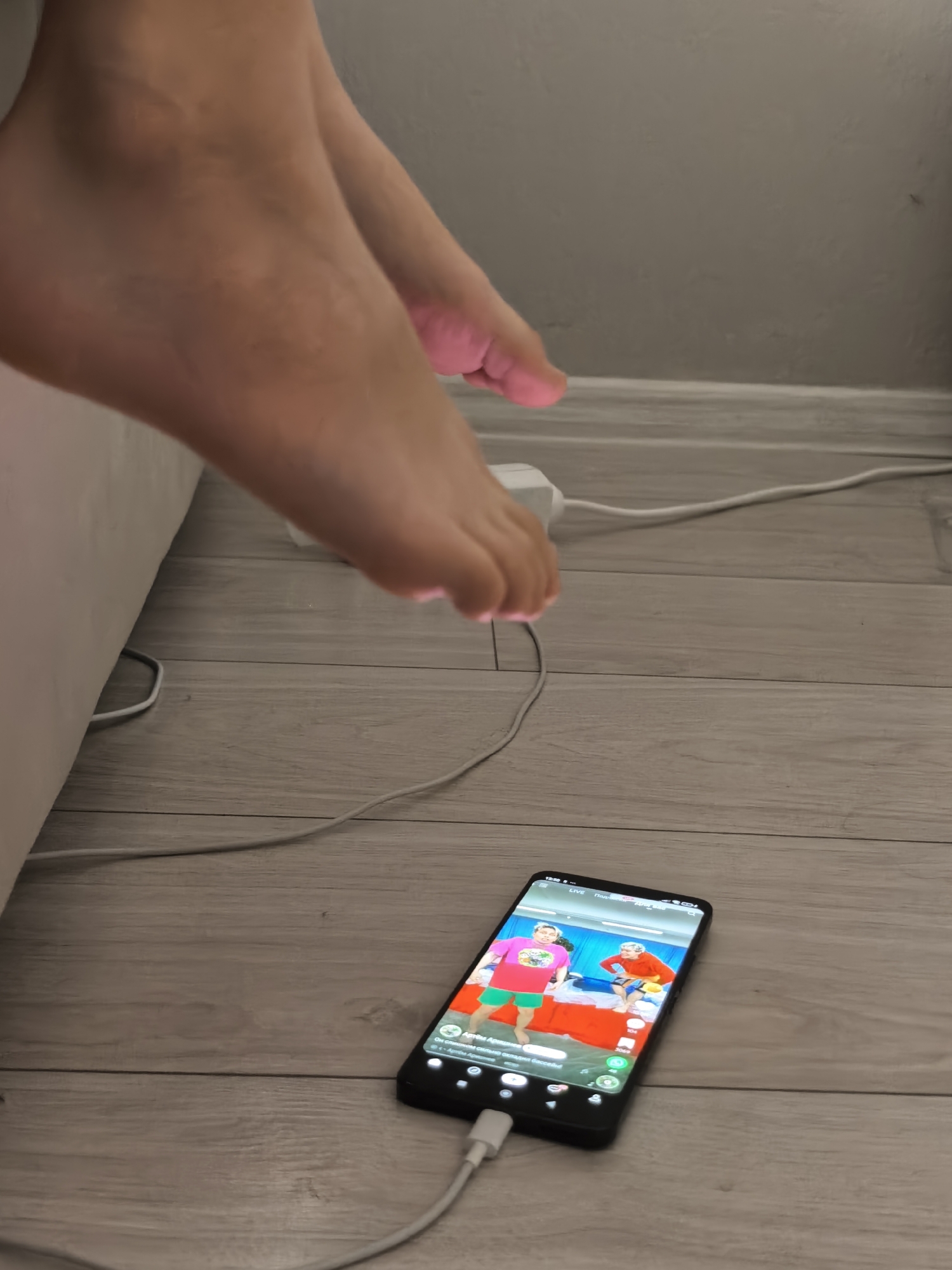 The image shows a person's bare feet and a smartphone placed on a wooden floor. The smartphone is playing a video that appears to be a live stream, with two people visible on the screen. A charging cable is connected to the smartphone. The background consists of a wall and a white object that might be a bed or couch. The scene is indoors and appears to be a domestic setting. There is no indication of the specific location within a city.