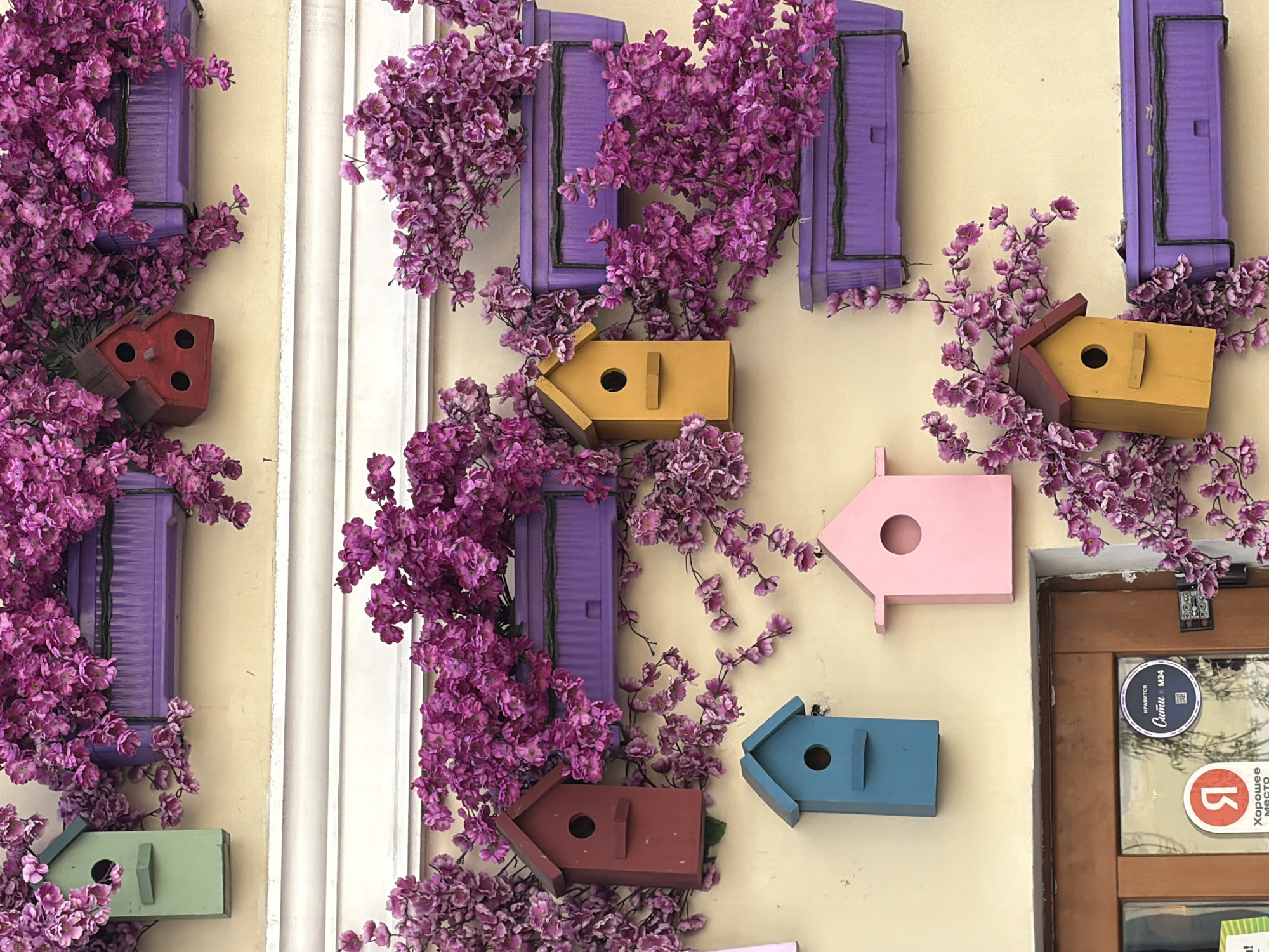The image shows a wall in Moscow, Russia, adorned with colorful birdhouses and purple flowers. The birdhouses are painted in various colors, including red, yellow, pink, blue, and green. The purple flowers are arranged around the birdhouses and windows. The wall is painted in a light yellow color. A doorway is visible on the right side of the image, with stickers on its glass window.