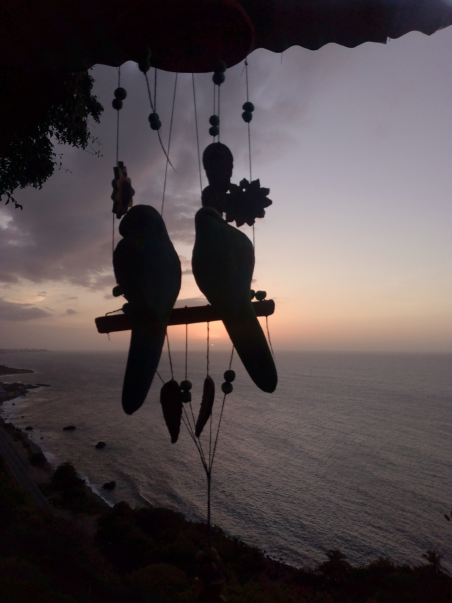 A low-angle, full shot captures a silhouetted wind chime against a dusky sky over the ocean. The wind chime, suspended from an unseen structure, features two dark, parrot-shaped ornaments perched on a wooden dowel, with several smaller, dark, pod-shaped ornaments dangling below. The backdrop is a serene seascape with a calm ocean reflecting the soft hues of the twilight sky, a faint sliver of the setting sun visible on the horizon. The coastline, with dark trees and rocky outcrops, is visible in the distance on the left. The scene is devoid of people and their actions.