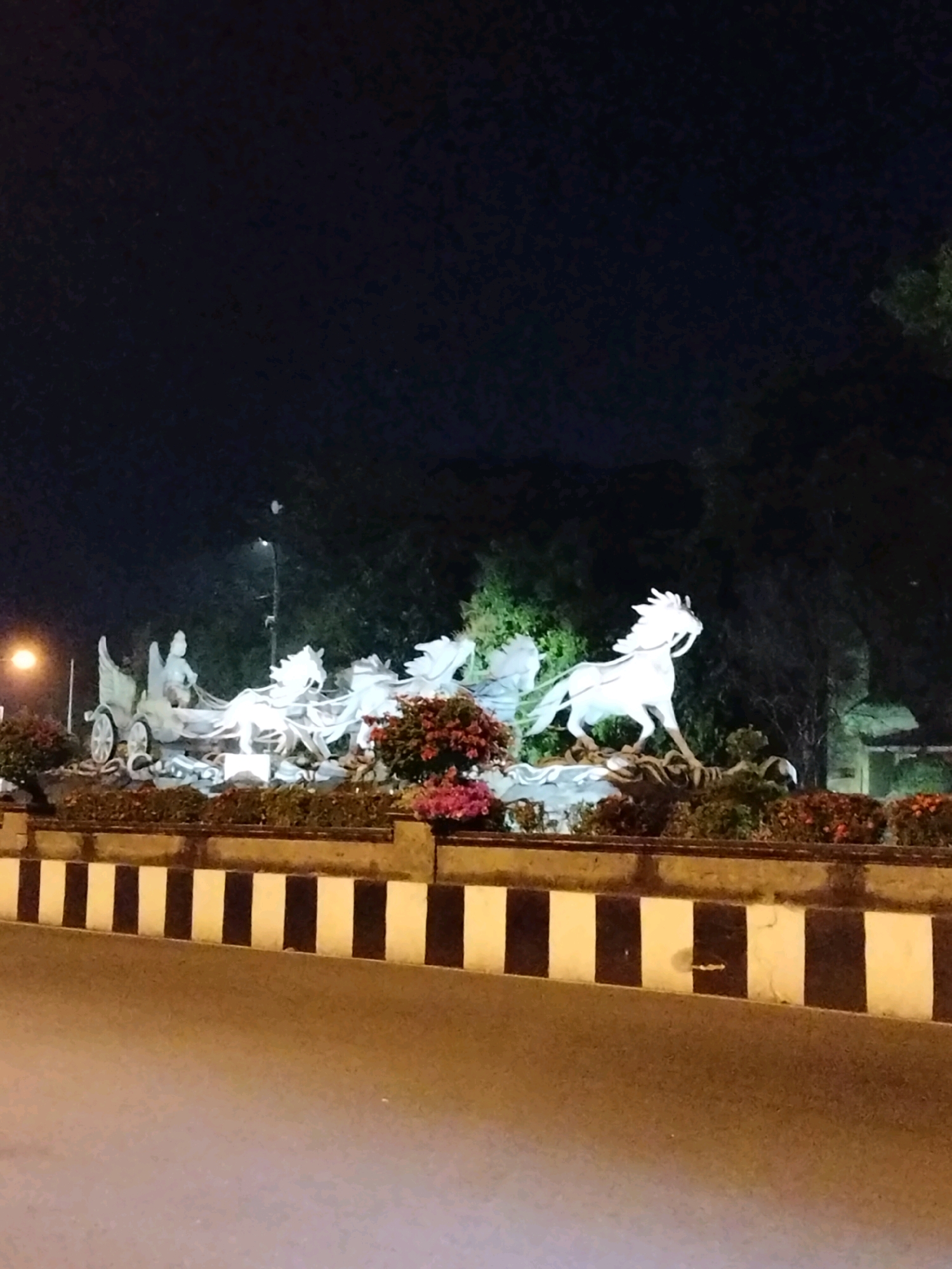 A nighttime, low-angle, medium shot captures a white sculpture of a chariot with four horses, depicting a mythological scene, situated in a roundabout in the city of Purworejo, Indonesia. The sculpture, illuminated by artificial lights, is adorned with floral arrangements in shades of pink and red along its base. A traffic island with black and white striped markings separates the roundabout from the road. The background is dark, with silhouetted trees and a street lamp visible. The scene is captured with a slight blur, indicating motion or a handheld camera.