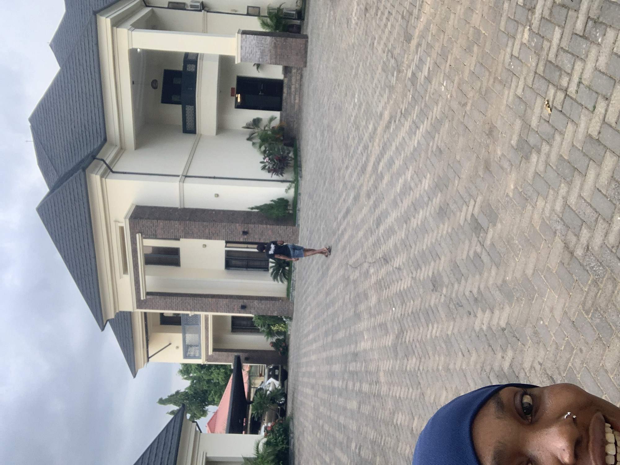 A person in a blue headscarf takes a selfie in the foreground, looking slightly to the right. Behind them, a paved driveway leads to a large, modern, cream-colored house with a dark gray, multi-pitched roof. The house features dark windows, balconies with decorative railings, and brick accents on some pillars. A person wearing a black t-shirt and denim shorts stands in the middle ground, facing away from the camera and walking on the paved driveway. To the left, a covered parking area shelters a car, with lush green plants visible around it. The scene is set under an overcast sky, typical of the weather in Abuja, Nigeria.